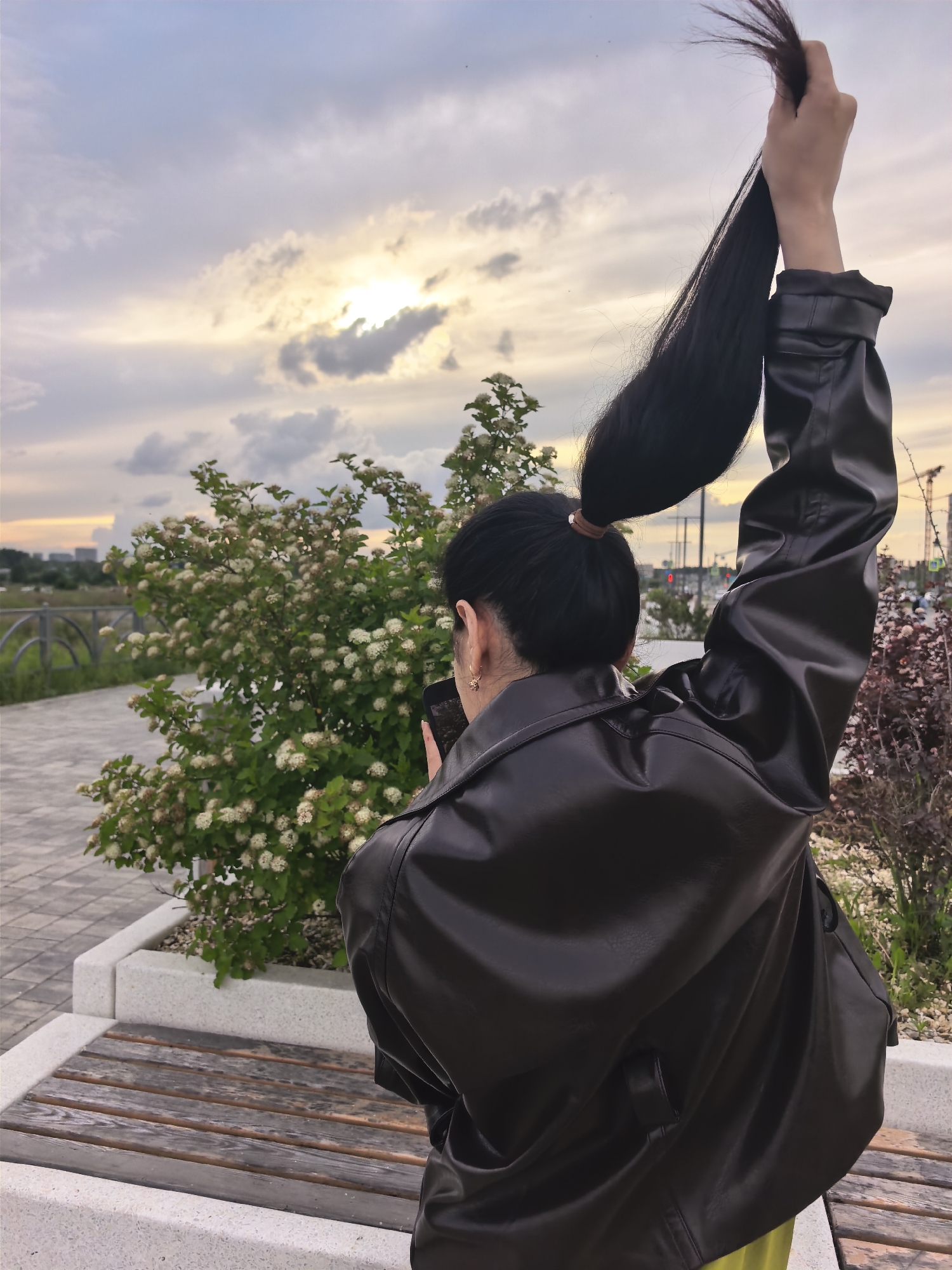 A person with dark hair pulled into a high ponytail, wearing a black leather jacket and a bright yellow garment, stands facing away from the camera, holding their hair up in their right hand. The person is holding a dark-colored mobile phone in their left hand, angled towards a flowering bush. They are positioned on a paved walkway, with a wooden bench in the foreground and a modern planter with a small tree or shrub. In the background, a cloudy sky with the sun peeking through can be seen, along with distant city buildings and construction cranes. The lighting suggests it is either early morning or late evening.