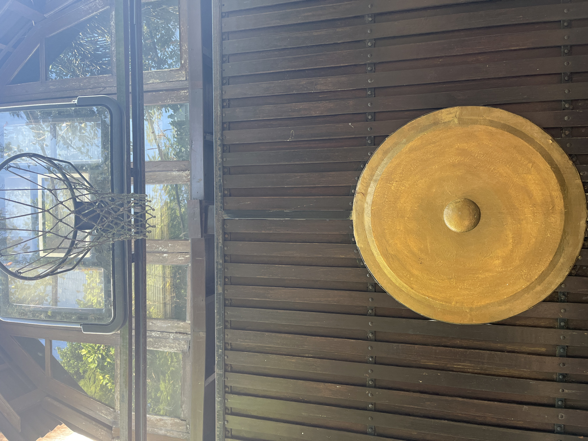 The image shows a basketball hoop mounted on a wooden structure with a large golden gong below it. The basketball hoop has a transparent backboard and a metal rim with a net. The wooden structure appears to be part of a building, with windows reflecting trees and the sky. The gong is circular with a raised dome in the center and is attached to the wooden structure. The background is a vertical wooden fence with visible rivets. The scene is shot from a low angle, looking upwards towards the basketball hoop. The overall setting suggests an outdoor recreational area within a Kuta Selatan, Indonesia, property.