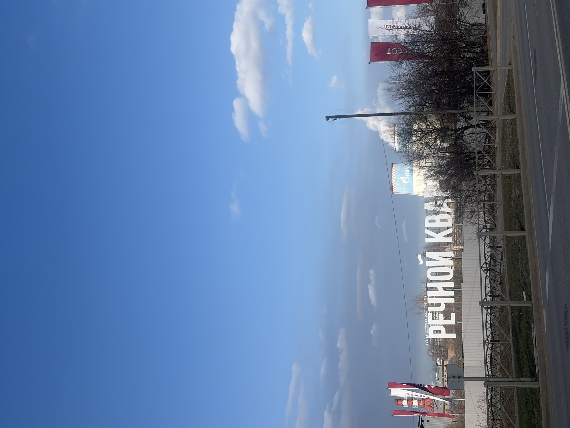 The image depicts an industrial and development area in Saint Petersburg, Russia, identifiable by the Pravoberezhnaya Thermal Power Plant (CHPP-26 / ТЭЦ-26) featuring cooling towers with the Gazprom logo and red-and-white striped chimneys. A large sign in the background reads "ТЭК ПРАВОБЕРЕЖНАЯ КОТЕЛЬНАЯ." In the midground, prominent white dimensional letters spell "РЕЧНОЙ КВА" (likely part of "РЕЧНОЙ КВАРТАЛ" or River Quarter), positioned behind a light-colored fence. To the left and right of these letters, several tall red and white vertical banners are visible, some with Cyrillic text. A line of bare-branched trees and bushes runs across the midground. In the foreground, an asphalt road surface with a white line is visible, separated from a patch of sparse, dry ground by a dark grey metal barrier fence with a geometric pattern. A dark utility pole stands on the right side. The sky above is largely clear blue with scattered white cumulus clouds. No people or vehicles are present.