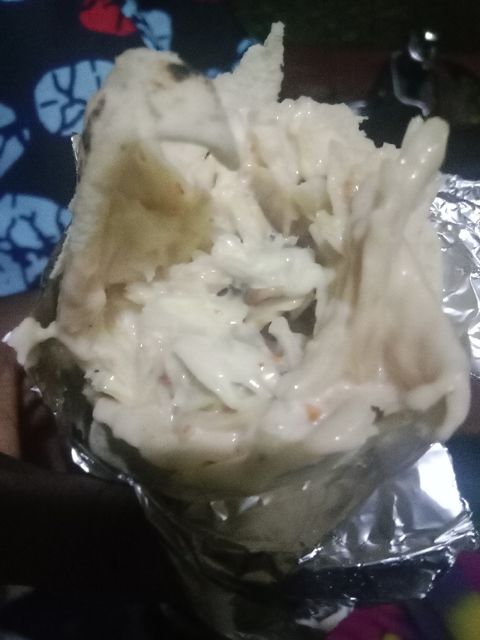 A partially eaten shawarma wrap rests in foil, exposing its filling. Creamy white sauce liberally coats shredded lettuce, and occasional bits of shredded carrots. The wrap itself appears to be a light tan color with darker spots. A colorful patterned fabric is visible behind the shawarma, partially obscured by a hand holding the wrap.