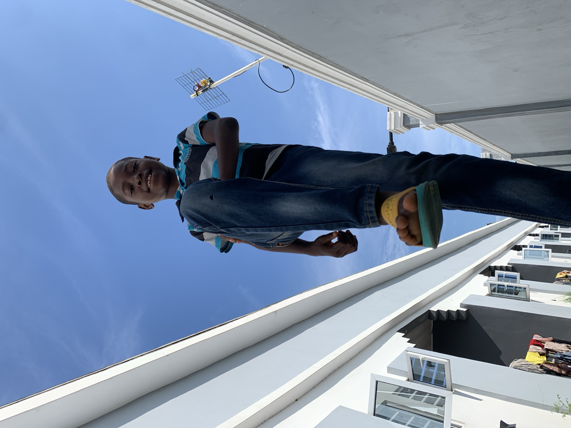 This low-angle, full-body shot captures a young boy standing outdoors against a clear blue sky. The boy, who appears to be of African descent, is smiling broadly at the camera. He is dressed in a casual striped t-shirt with blue, black, and white horizontal bands, and blue denim jeans, which show a small tear on the right knee. He wears yellow sandals with teal-colored soles on his feet.
The unique perspective, looking upwards, makes the boy appear tall and dominant in the frame, with architectural elements forming the background. To the right of the boy, a multi-story building with a modern design is visible, featuring white and grey facades, multiple balconies or ledges, and open windows. An old-fashioned grid-style television antenna is mounted on a pole, extending into the clear sky near the top left of the boy's head.
The sky is a vivid, cloudless blue, indicating a bright and sunny day, likely morning or afternoon. The boy's joyful expression conveys happiness. The setting is an urban or semi-urban environment, consistent with Ibasa, Nigeria. No discernible text is visible on any of the subjects or in the background.