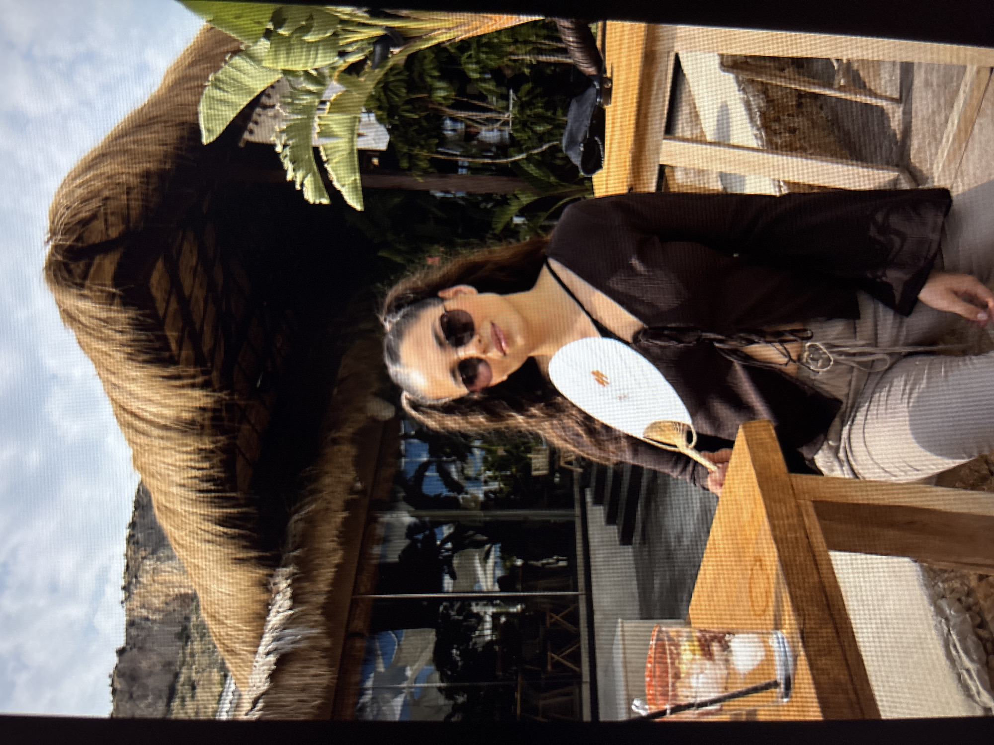 A young woman with dark hair in pigtails and sunglasses holds a white fan. She is wearing a dark, long-sleeved top with a tie closure and light-colored pants. She is seated at a wooden table outdoors, with a glass of iced beverage to her left. In the background, there is a thatched-roof structure, large green plants, and a cliff face. The setting appears to be a restaurant or outdoor venue. The scene is well-lit, suggesting it is daytime. There is no information in the image that indicates the location is in Sofia, Bulgaria.
