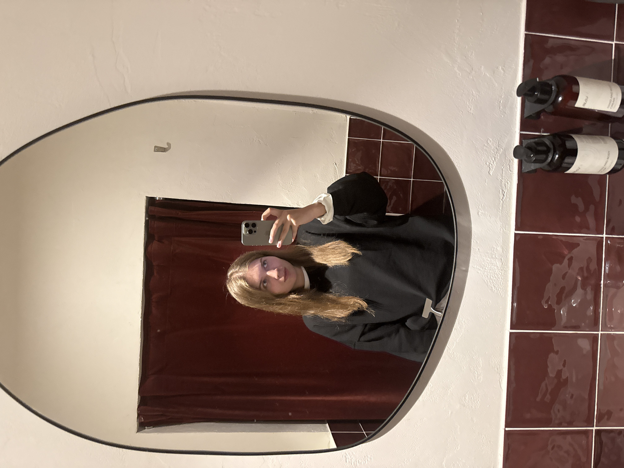 A young woman with light brown hair is reflected in an oval mirror in what appears to be a bathroom. She is holding a smartphone up and angled towards herself, presumably taking a picture. She is wearing a dark sweatshirt with white cuffs. The background of the mirror reflects a dark red curtain or tapestry. To the right of the mirror, on the wall, are dark red square tiles and two bottles of liquid soap mounted on the wall. The bathroom has white walls. The scene appears to be indoors. The exact location in Belgrade, Serbia, is impossible to determine from the provided image.