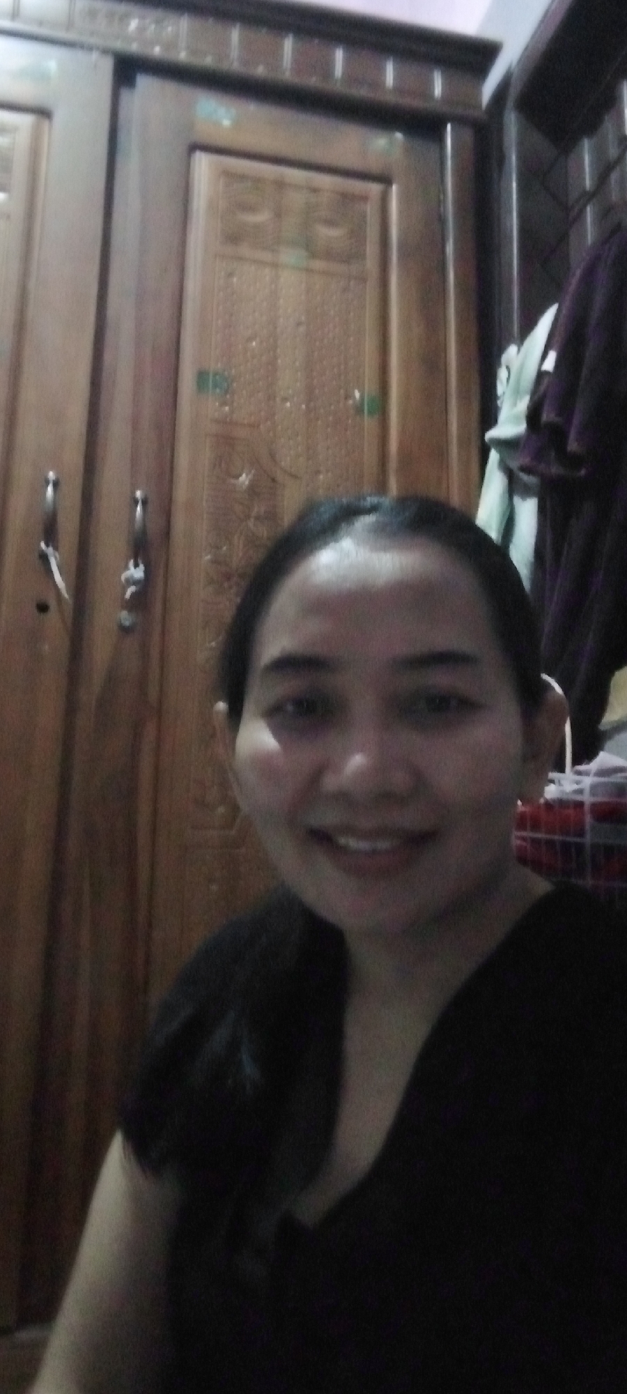 A selfie is taken by a woman with dark hair and fair skin in front of a wooden wardrobe. The wardrobe has two doors with decorative carvings and silver handles. The woman is wearing a black shirt and smiling. The background is dimly lit. The scene is indoors, likely a bedroom.