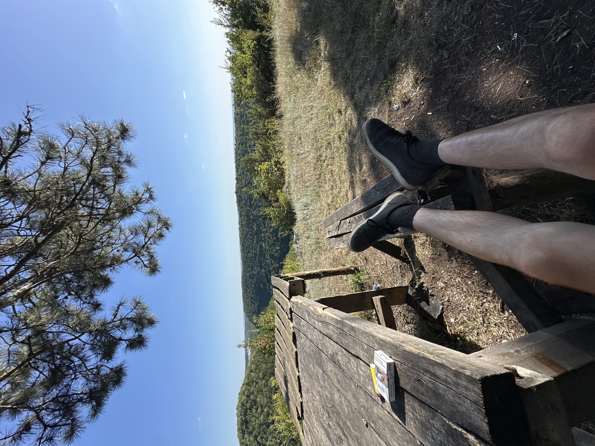 The image depicts a man's legs and feet resting on a wooden bench, with a pack of cigarettes and a lighter on a wooden picnic table to his left. The setting appears to be a scenic overlook with a panoramic view of a lush, green valley and rolling hills under a clear blue sky. Pine branches hang overhead, casting dappled shadows. The environment suggests a rural or natural area, possibly a park or a viewpoint overlooking a natural landscape near Pryvorottia Druhe, Ukraine. The scene is peaceful and suggests a moment of relaxation or a break during an outdoor activity.