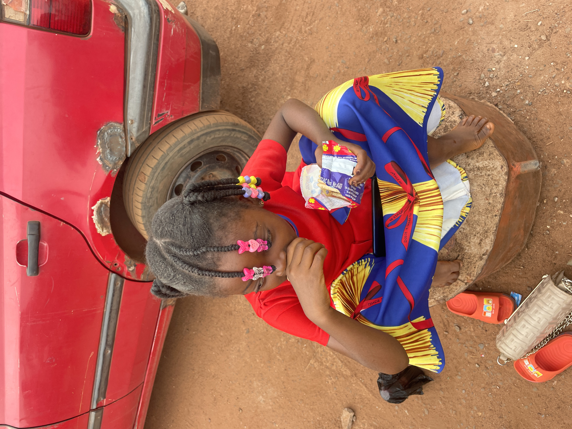 A young girl with braided hair sits on a tree stump on a dirt ground in Otukpa, Nigeria. She wears a red t-shirt and a brightly colored dress in shades of yellow, blue, and red. Pink heart-shaped clips adorn her hair. She holds a snack in her hand, looking downward. Nearby are a pair of orange sandals and a white purse with a chain. A red car with visible bodywork and a visible tire is partially in the shot.