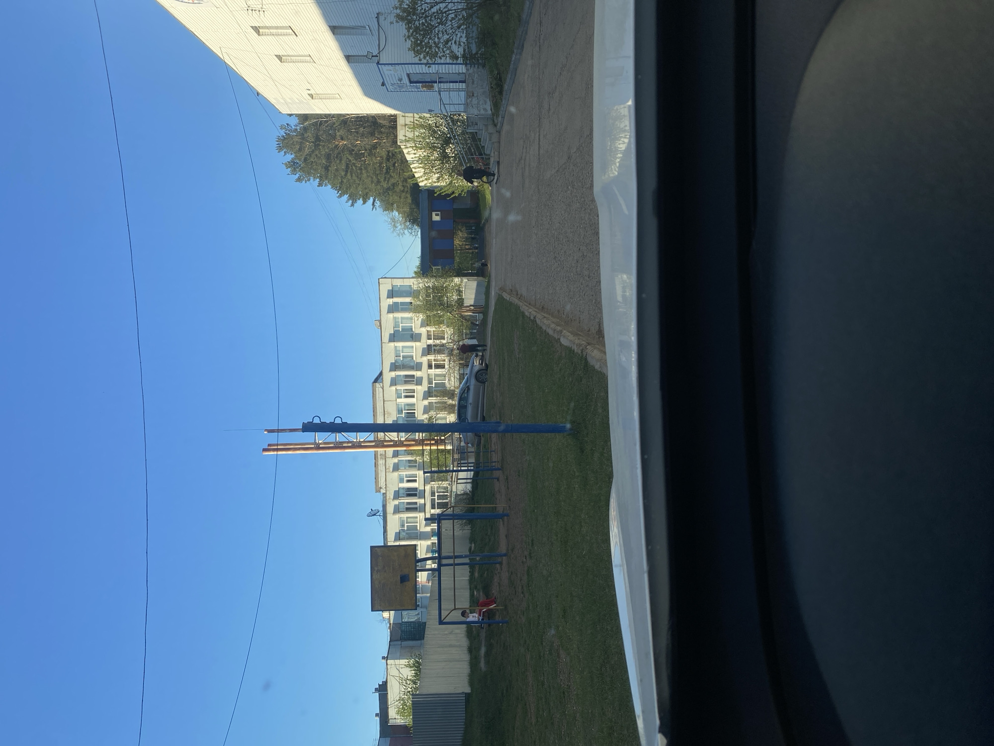 The image, taken in the city of Chaikovsky, Russia, depicts a view from inside a vehicle looking out onto a residential area. A playground with blue metal structures is visible, including a basketball hoop and swing set where two children sit. Behind the playground, multiple multi-story apartment buildings stand, characterized by white facades and numerous windows. The sky is clear and blue. A utility pole with attached hardware is visible near the playground. A paved walkway runs along the side of the frame. The interior of the vehicle is dark.