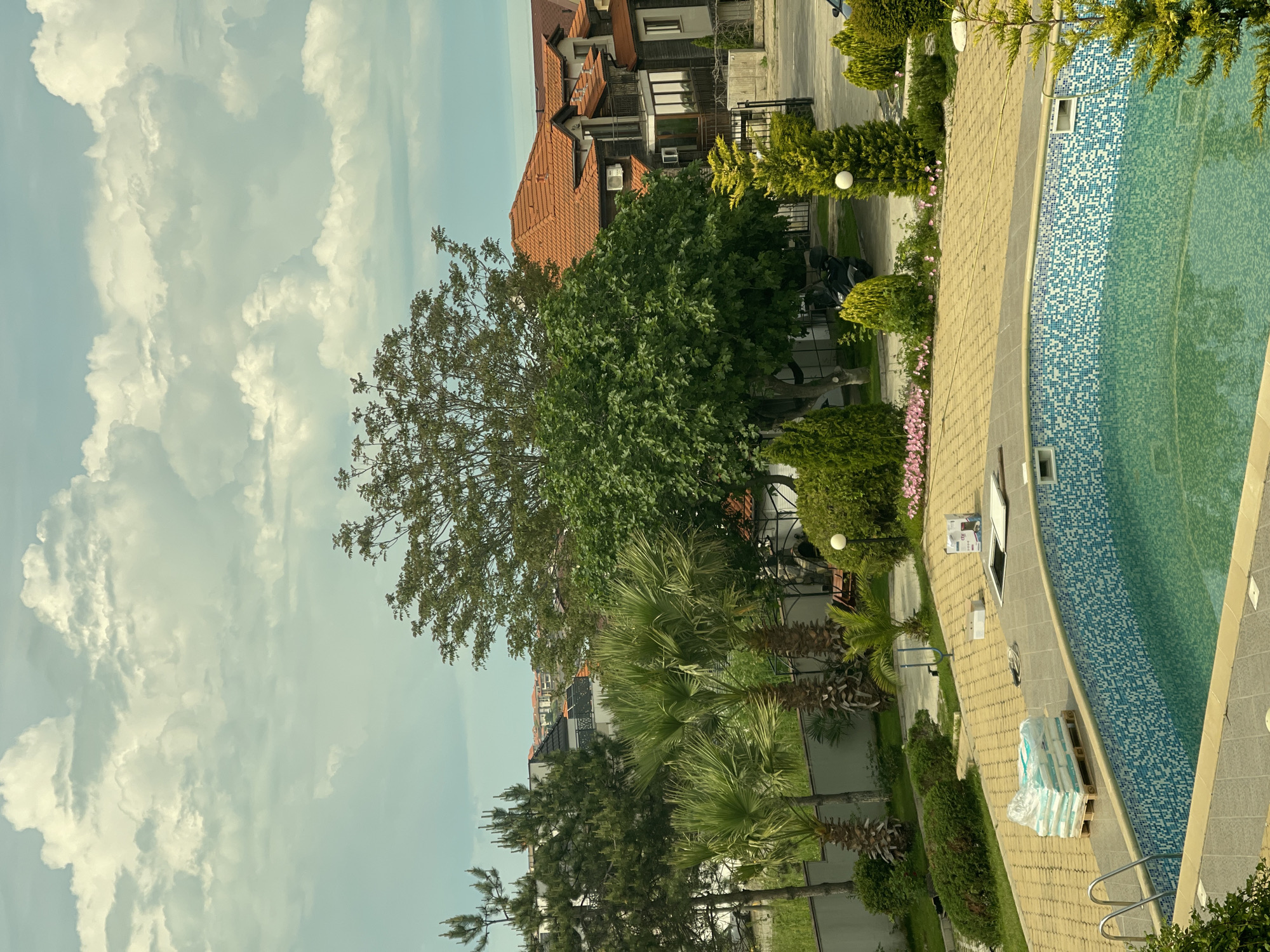 The image presents a scene in Sozopol, Bulgaria, capturing a view of the sky, buildings, trees, a pool, and a walkway. The sky is filled with fluffy, white clouds. A building with a brown, tiled roof is visible, along with other structures. Lush green trees, including palm trees, dominate the landscape. A pool with blue and white mosaic tiles is situated next to a paved walkway. A stack of materials wrapped in plastic sits on the walkway near the pool's edge. The scene appears tranquil and residential.