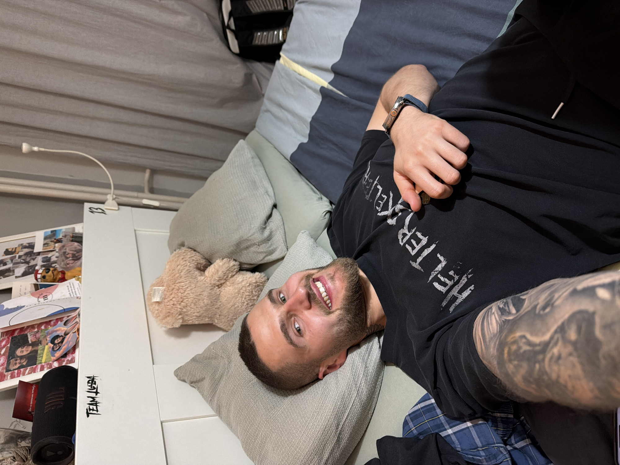 A young adult man, with short dark hair, a beard, and green eyes, lies smiling on a bed in what appears to be a bedroom in Sofia, Bulgaria. He is looking directly at the camera with a wide, happy smile, showing his teeth. He is wearing a black t-shirt with partially visible white text that reads "HELTER" and blue plaid pajama-style pants. A prominent, intricate tattoo covers his left arm. On his right wrist, he wears a watch with a blue band.
He is propped up by a light grey pillow, and another matching pillow is visible behind him. A small, light brown teddy bear is placed next to his head on the bed. The bedding features shades of light grey, dark grey, and blue. To the left of the bed, a white headboard or bed frame is visible, with various photos, cards, and what looks like magazine clippings attached. A black portable speaker or device rests on a surface next to these items. In the background, a flexible white clip-on lamp is attached to a pole or bed structure. Part of a black and white backpack is also visible at the top of the image.
Visible text includes "HELTER" on his t-shirt, "TEAMLEBAH" written vertically on the white bed frame, and the number "13" next to it. The scene conveys a relaxed and personal moment.