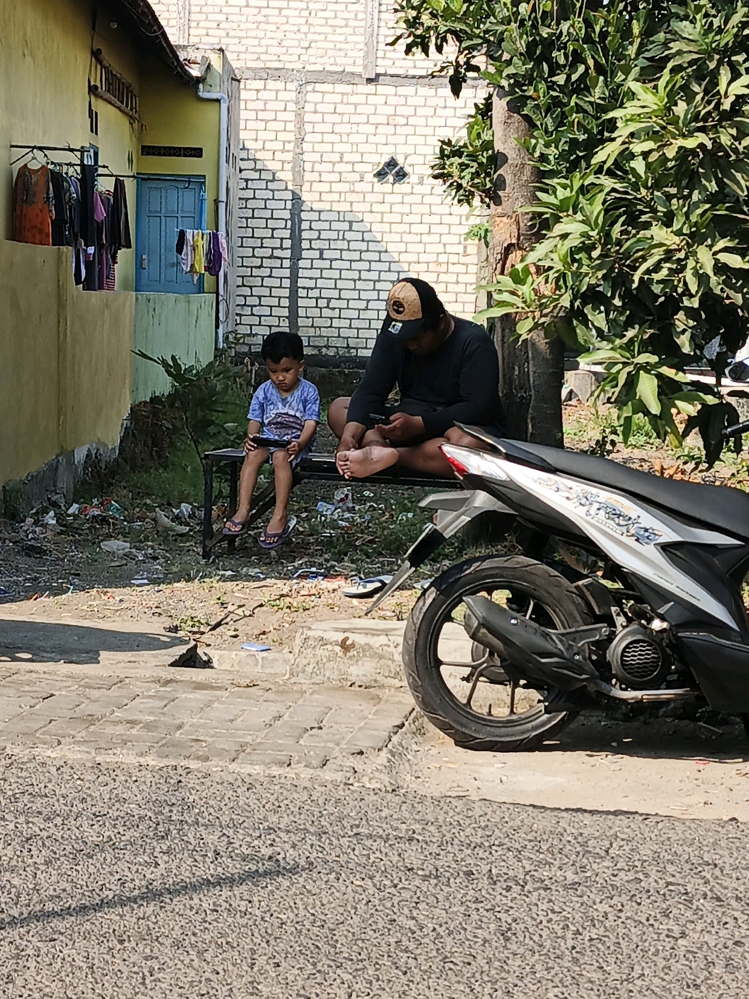Two individuals are seated on a bench outdoors in Boro, Indonesia. A young boy, wearing a blue t-shirt and shorts, is looking at a mobile phone he holds in his hands. Beside him, an adult, wearing a black long-sleeved shirt and a baseball cap, is also looking at a mobile phone. They are positioned on a bench situated in front of a building with a yellow facade and a blue door. The ground around them is covered with debris and some vegetation. A parked motorcycle is visible in the foreground, to the right of the bench. The background shows a brick building and a tree with lush green leaves. The scene is bathed in sunlight, casting shadows on the ground.