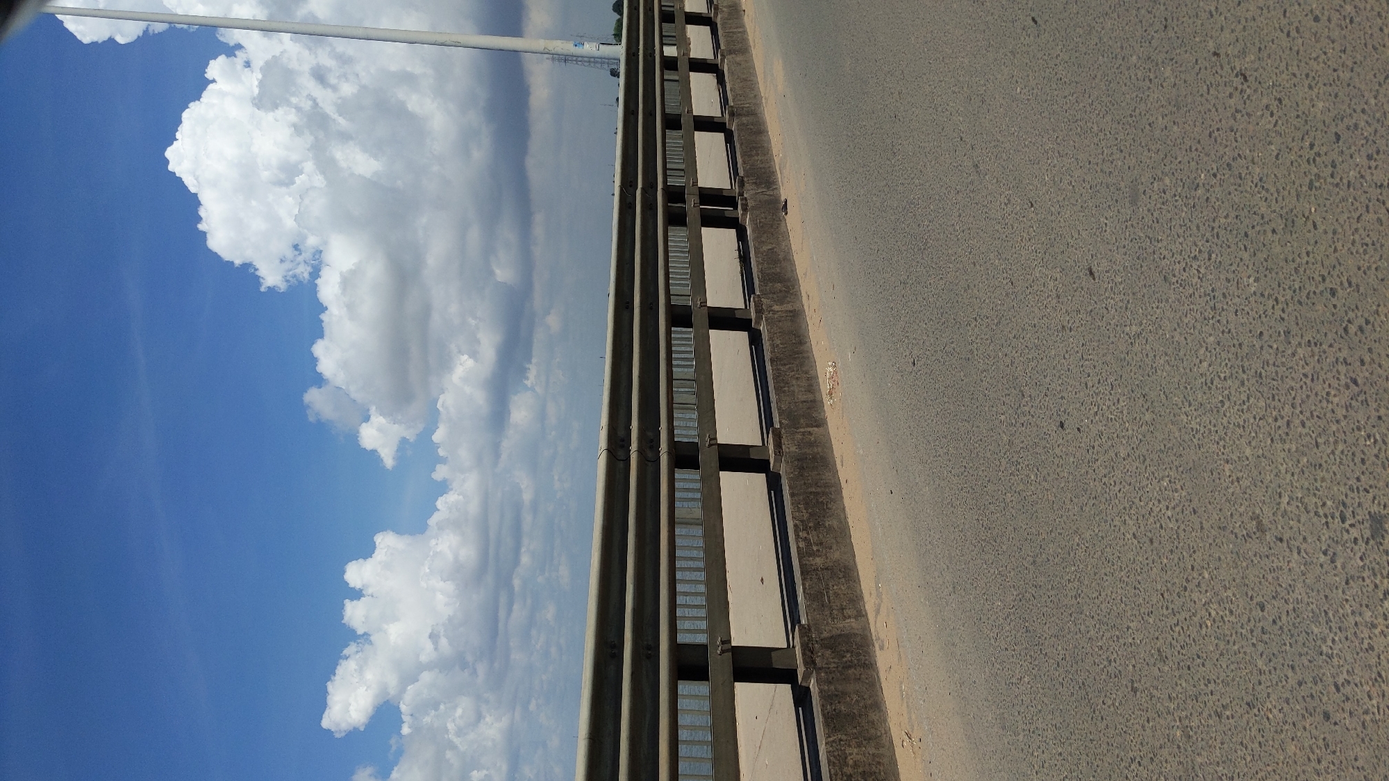 The image shows a bridge in Dar es-Salaam, Tanzania. The sky is blue with white clouds. The bridge has a gray concrete barrier on the left. The road surface on the bridge is gray asphalt.