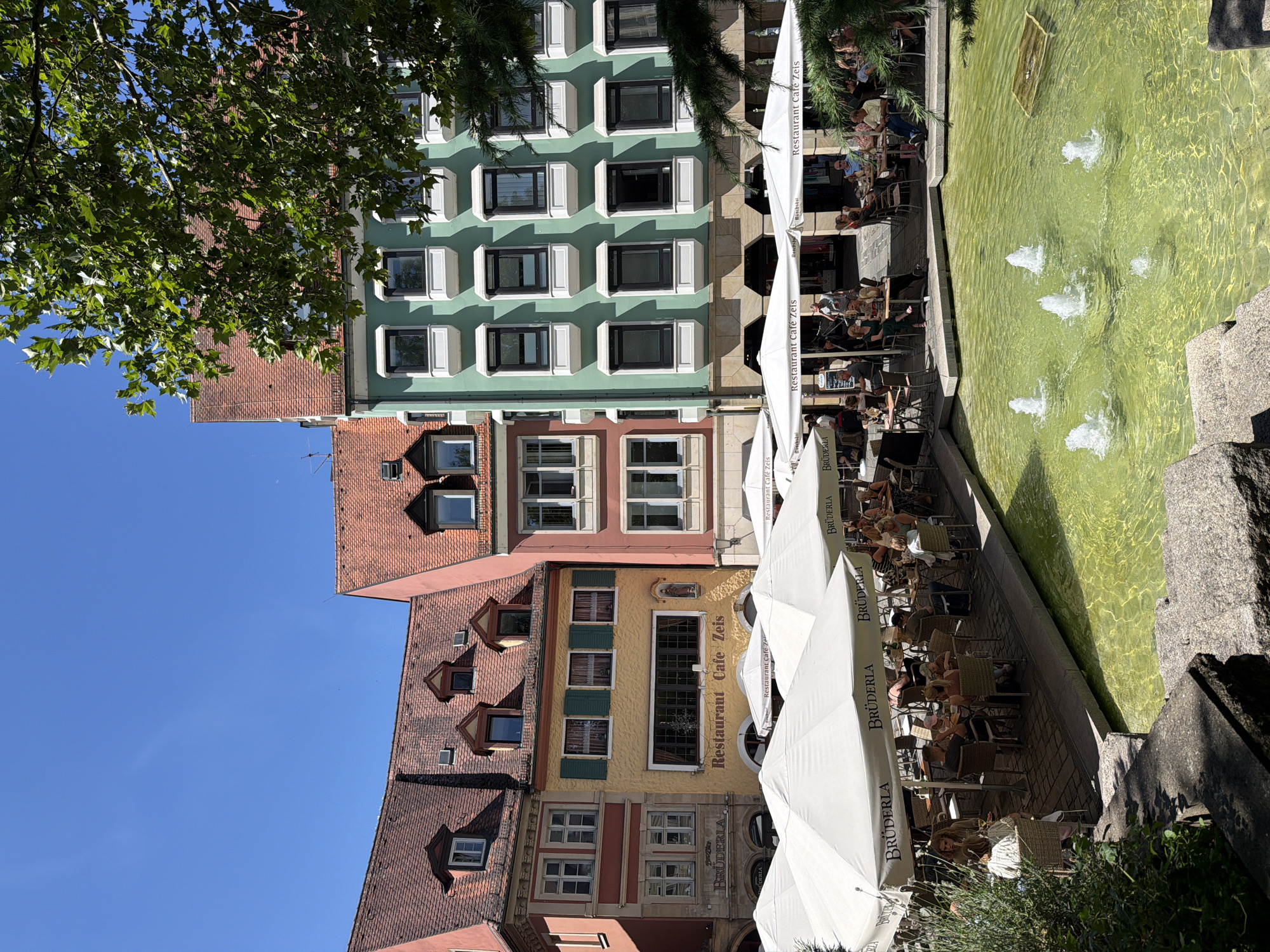 This image depicts a sunny outdoor seating area of a restaurant called "Restaurant Cafe Zeis" in Bamberg, Germany. The restaurant is situated in a square with buildings characterized by their traditional German architecture, featuring red tile roofs and colorful facades, including a prominent yellow building with green shutters. White umbrellas provide shade for tables occupied by patrons enjoying their meals and drinks. In the foreground, a fountain with multiple jets sprays water into a bright green pool, reflecting the clear blue sky. Several people are visible seated at tables, engaged in conversation or dining. Lush green trees and foliage frame the scene on the right side of the image. The ground is paved with interlocking bricks.