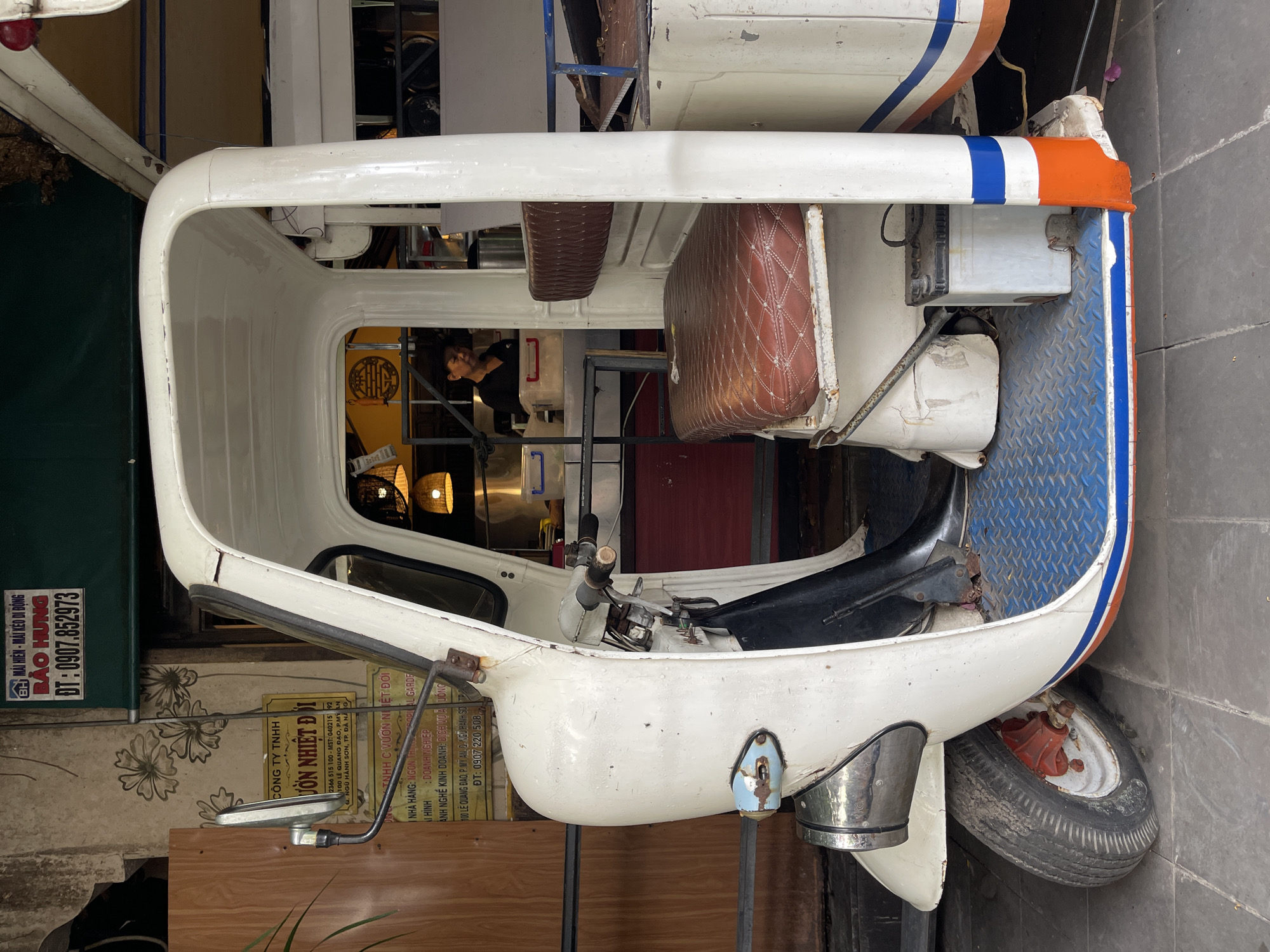 The image captures a three-wheeled vehicle, possibly a tuk-tuk or a similar motor-rickshaw, parked on a tiled surface in what appears to be a street or an alleyway in Da Nang, Vietnam. The vehicle is predominantly white with some red and blue accents. It has a driver's seat and a passenger seat upholstered in brown, diamond-patterned material. A battery is visible near the passenger seat. Behind the vehicle, a woman is partially visible, wearing a dark top and standing near a counter or serving area, suggesting it might be a food or beverage establishment. Signs with Vietnamese text are displayed on the wall behind the vehicle, indicating business information. The overall scene suggests a commercial area, possibly with a focus on street food or local services.