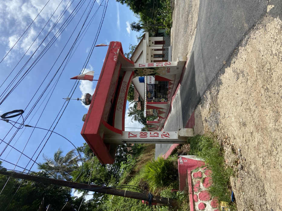 The image shows a red and white archway with the number 17084579 on the left and 17082179 on the right. The archway is located on a paved road that leads up a hill. On the left side of the road there is a low retaining wall with red and white decorative stones and a "GENTENG" sign. On the right side of the road, there are two buildings. The closer one is painted white with a red roof and has a sign with "GGSP" written on it. The further building has a similar style. There is a small Indonesian flag attached to the archway, and a larger Indonesian flag is attached to the wall of the closer building. The sky is blue with white clouds, and there is a lot of green vegetation surrounding the road. The scene is located in Cipatat, Indonesia, at latitude -6.778 and longitude 107.437. There are no people in the scene. The archway is likely a gateway to a village or community. The scene is taken during the day.