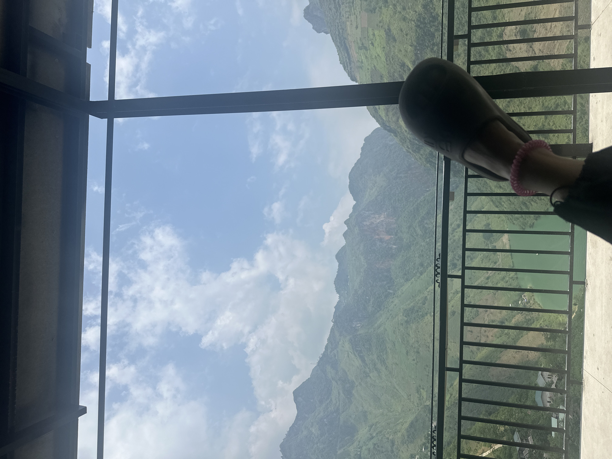 The image presents a view from a structure with dark metal framing and railings. Outside, there's a mountainous landscape, predominantly green with vegetation. A body of water, also green, is visible behind the railings. A part of a person's leg and foot, wearing a sandal and a pink scrunchie-like accessory, is in the foreground, partially obscuring the view. The sky is blue with scattered clouds. The location is likely Mèo Vạc Commune, Vietnam, based on the characteristic mountainous terrain.