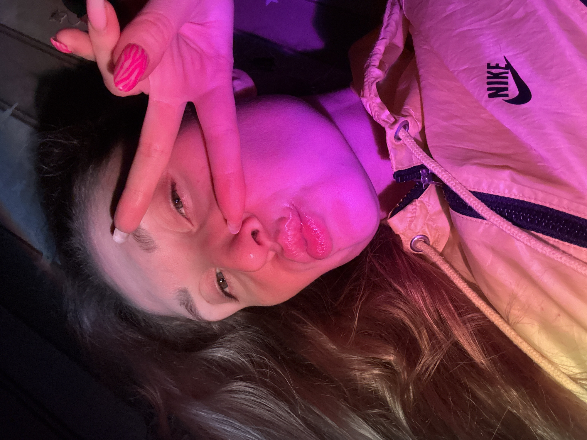 A young woman with long, dark blonde hair is lying down, angled towards the camera. She is making a peace sign with her right hand and has pink nail polish with a zebra print design on one nail. She is wearing a yellow Nike jacket and has a purple light shining on her face. The background is dark. It's impossible to determine that it was taken in the city of Chaikovsky, Russia.