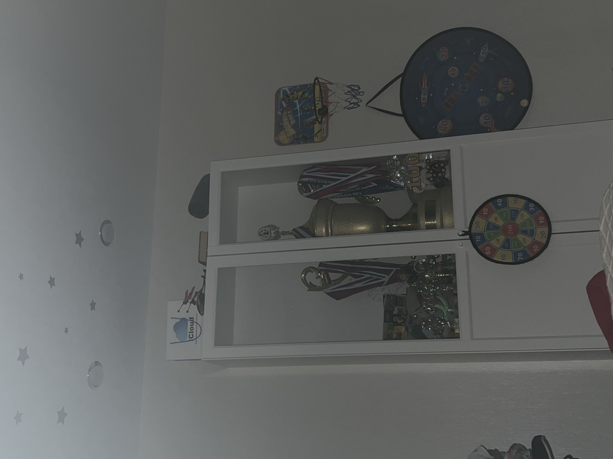 The image depicts an indoor scene, likely a child's bedroom, filled with various items related to sports and achievement. The main focus is a white display cabinet filled with trophies and medals, suggesting a history of athletic accomplishments. To the right of the cabinet, a small basketball hoop with a colorful backboard is mounted on the wall, and below it, a circular dartboard hangs, also featuring colorful numbers and graphics. On the wall above the cabinet, star-shaped decals are visible, further contributing to a youthful atmosphere. The overall impression is of a room decorated to celebrate achievements and encourage play. The image does not contain any specific indicators related to the city of Volgograd, Russia.