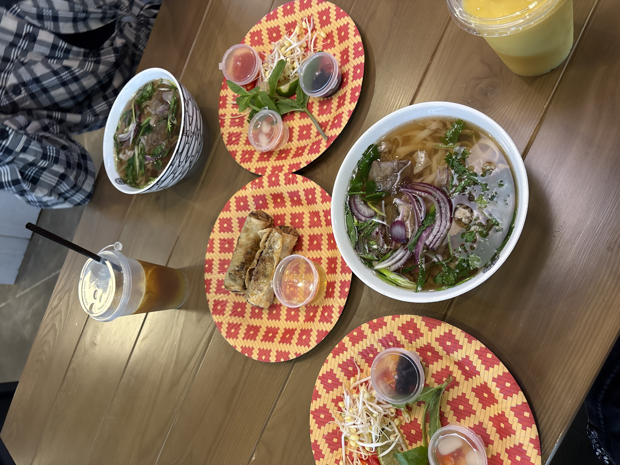 This image captures a close-up view of a meal served on a wooden table in an indoor restaurant setting in Krasnoyarsk, Russia. The specific address within Krasnoyarsk is not discernible from the visual information provided. **People:** Two individuals are partially visible. In the upper-right quadrant, a person's torso and arms are seen, wearing a black and white plaid long-sleeved shirt. In the lower-left corner, a portion of a leg or lap, clad in dark fabric, is visible. **Subjects/Objects:** The wooden table, light brown with prominent grain, is laden with Vietnamese cuisine and beverages. * **Main Dishes:** Two bowls of pho are present. One in the upper-center is a white bowl with a black geometric pattern around its exterior, filled with a clear broth, flat rice noodles, slices of beef, red onion, and green herbs (likely cilantro and scallions). A second similar bowl of pho is positioned in the lower-center. * **Appetizers:** On a circular red and yellow woven placemat in the upper-right, three golden-brown fried spring rolls are arranged, accompanied by a small clear plastic cup containing an amber-colored dipping sauce. * **Garnishes:** Two additional circular red and yellow woven placemats hold various garnishes. * The placemat in the lower-left displays a pile of bean sprouts, a lime wedge, mint leaves, and a few red chili slices, alongside two small clear plastic cups of dipping sauces (one dark liquid, one lighter liquid with chili flakes). * The placemat in the mid-right shows a similar arrangement of bean sprouts, a lime wedge, and mint leaves, with three small clear plastic cups of dipping sauces (one red, one clear/white, and one dark liquid with green specks). * **Beverages:** * In the upper-left, a clear plastic cup with a lid and a black straw contains an iced, amber-colored beverage. The lid has "CAUTION: HOT!" printed on it, despite the presence of ice. * In the lower-right, another clear plastic cup with a lid contains a thick, yellow-orange liquid, resembling a smoothie or juice. **Actions/Interactions:** The arrangement of the dishes, garnishes, and drinks suggests that a meal is either being served or is in progress. The visible individuals are seated at the table, indicating they are the patrons consuming the meal. **Environment:** The background is largely out of focus. The upper-left shows a dark, indistinct surface, possibly a corner or another piece of furniture. The upper-right reveals a blurry light-colored wall or doorway. The