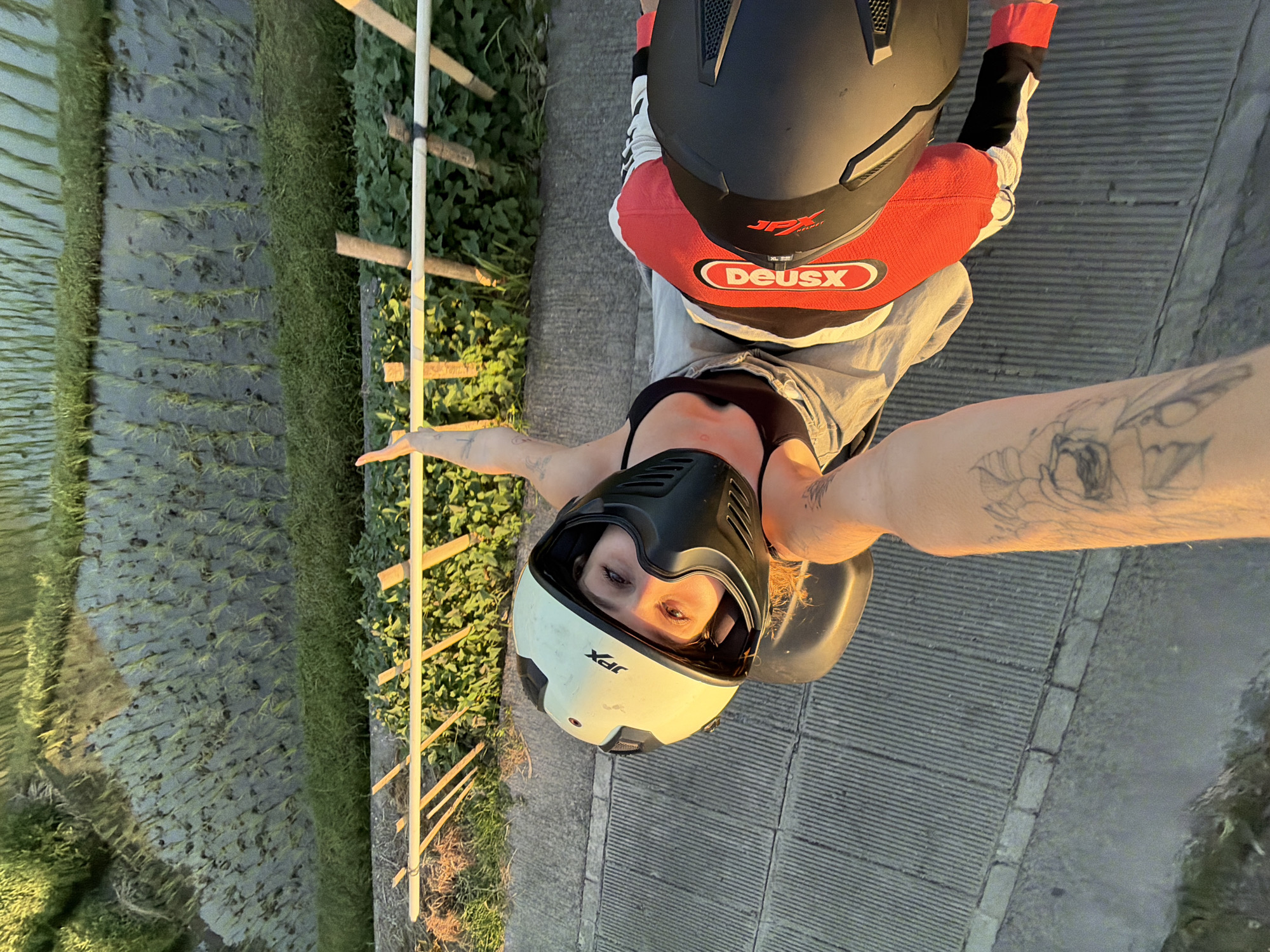 An individual wearing a white and black helmet and a black sports bra, with a red and black jacket is positioned on a scooter. The scooter is on a paved path adjacent to rice paddies. The individual is looking up towards the camera, their arm is extended upwards. A second individual's arm, with visible tattoos, is holding the scooter. The setting appears to be outdoors with natural lighting. The background shows rows of green rice shoots in flooded paddies, with a wooden fence along the edge. The overall scene is an outdoor activity in a rural or semi-rural area, possibly within North Kuta, Indonesia given the presence of rice paddies.