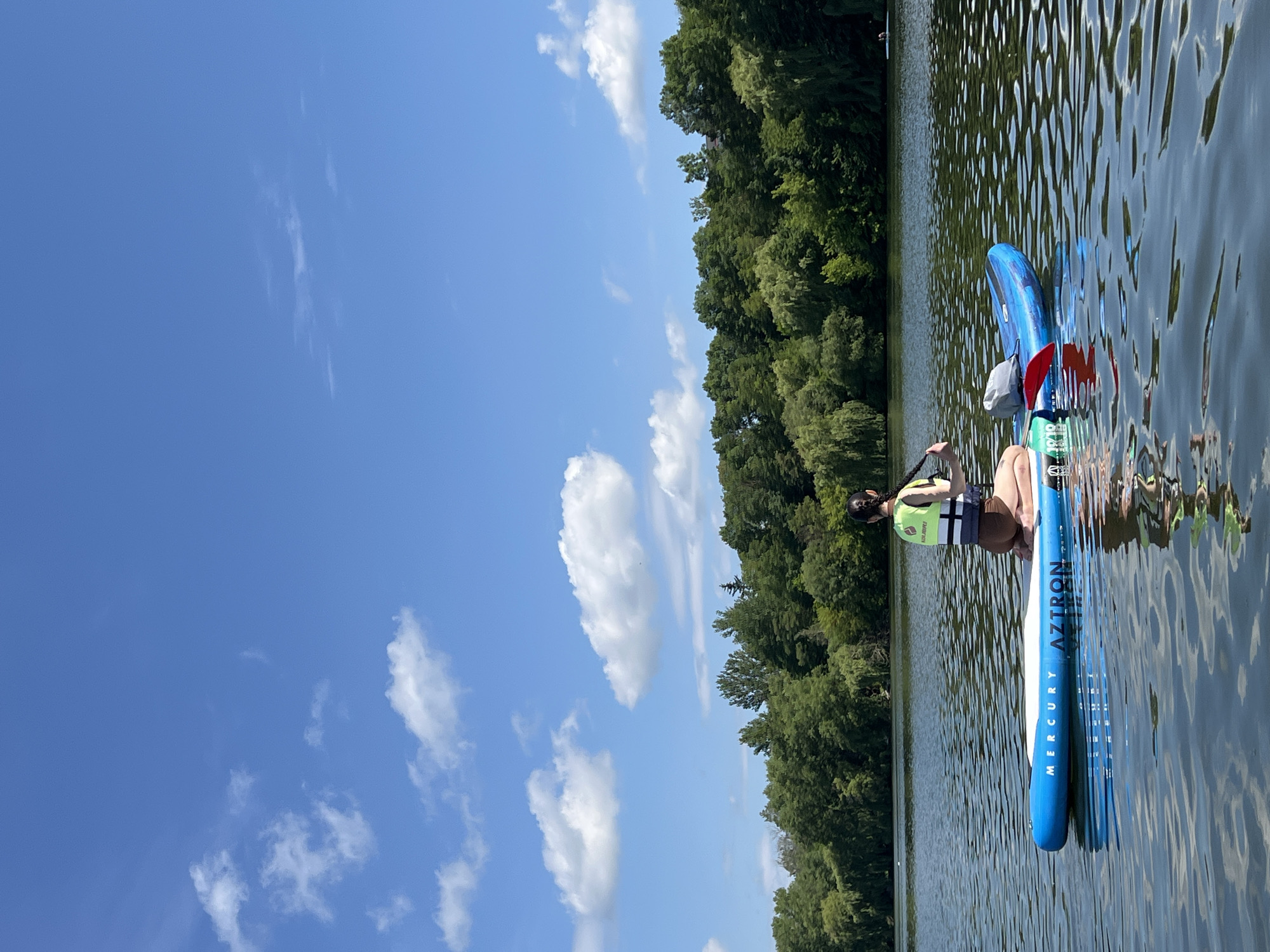 A person is paddleboarding on a calm lake with a line of trees in the background under a bright blue sky with scattered clouds. The person is kneeling on a blue and white paddleboard, wearing a green and white life vest. The paddleboard has the words "MERCURY AZTRON" printed on it. The water is rippled and reflects the sky and surrounding trees. In the distance, another person is visible on a small boat or kayak. The location is not in Ternopil, Ukraine.