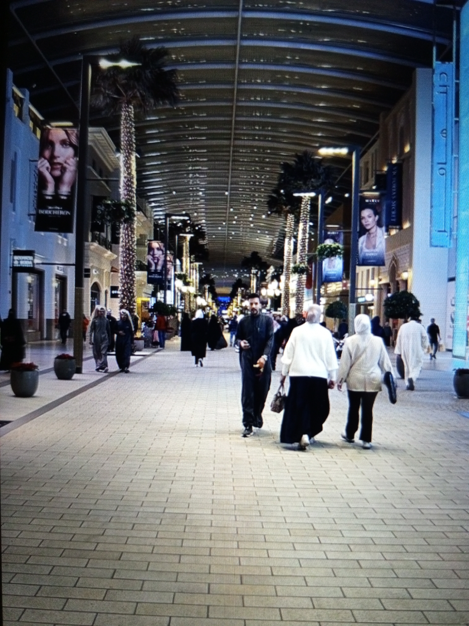 In the city of Golbembal, Nigeria, an indoor walkway is depicted, characterized by a high, arched ceiling with a modern, translucent covering that allows natural light to filter through. Palm trees, some adorned with festive lights, are strategically placed along the walkway, creating a tropical ambiance. The walkway is paved with light-colored rectangular tiles, arranged in a staggered pattern.
Several individuals are walking along the path. Two men in traditional attire, thobes and headscarves, are walking towards the viewer. Further down, a group of people are walking away from the camera. One woman is wearing a white abaya and black hijab, another is in a beige abaya and black hijab, and a third is in a white robe. A man in a dark abaya is walking towards the viewer, carrying a bag. Other figures are visible in the distance, suggesting a bustling environment.
Commercial signage is present, with banners featuring portraits of women. One banner displays a close-up of a woman's face, with the brand name "BOUCHERON" visible below. Another banner features a woman in a white top and light-colored skirt. The architecture suggests a high-end shopping or entertainment complex. The overall impression is one of a well-maintained and possibly luxurious public space.