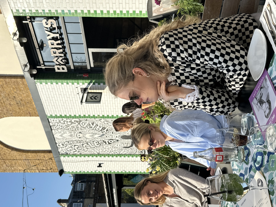 The image displays an outdoor cafe or restaurant seating area on a sunny day at Barry's Bootcamp, located on Wheler Street in Shoreditch, London, United Kingdom. Three blonde women are seated at a dark table with a patterned blue, green, and white tablecloth in the foreground. The woman on the right wears sunglasses and a black and white checkered long-sleeved cardigan with white crochet cuffs, her chin resting on her right hand. The central woman, also wearing sunglasses, has blonde hair and a light blue collared shirt, looking slightly to her left. The woman on the far left, with blonde hair, wears a grey collared shirt and is smiling towards the central woman.
On the table are multiple beverages, including a stemmed glass with a clear green cocktail containing mint leaves and ice, a transparent Coca-Cola bottle, and several other glasses with clear and dark liquids. A white square plate, cutlery (fork and knife), and various menus, including one titled "COCKTAIL LIST," are also present.
The background features the building facade, which is clad in white rectangular tiles with vertical green tiled stripes. A large black and white mural depicting abstract designs, an eye motif, and text partially visible as "WHELESTON YARDS" covers a section of the wall. Above an entrance, a metal sign with the name "BARRY'S" is visible. Planters containing green foliage and yellow flowers line the base of the wall. Above the tiled facade, a large arched structure of light brown brickwork is present, framed by a clear blue sky.