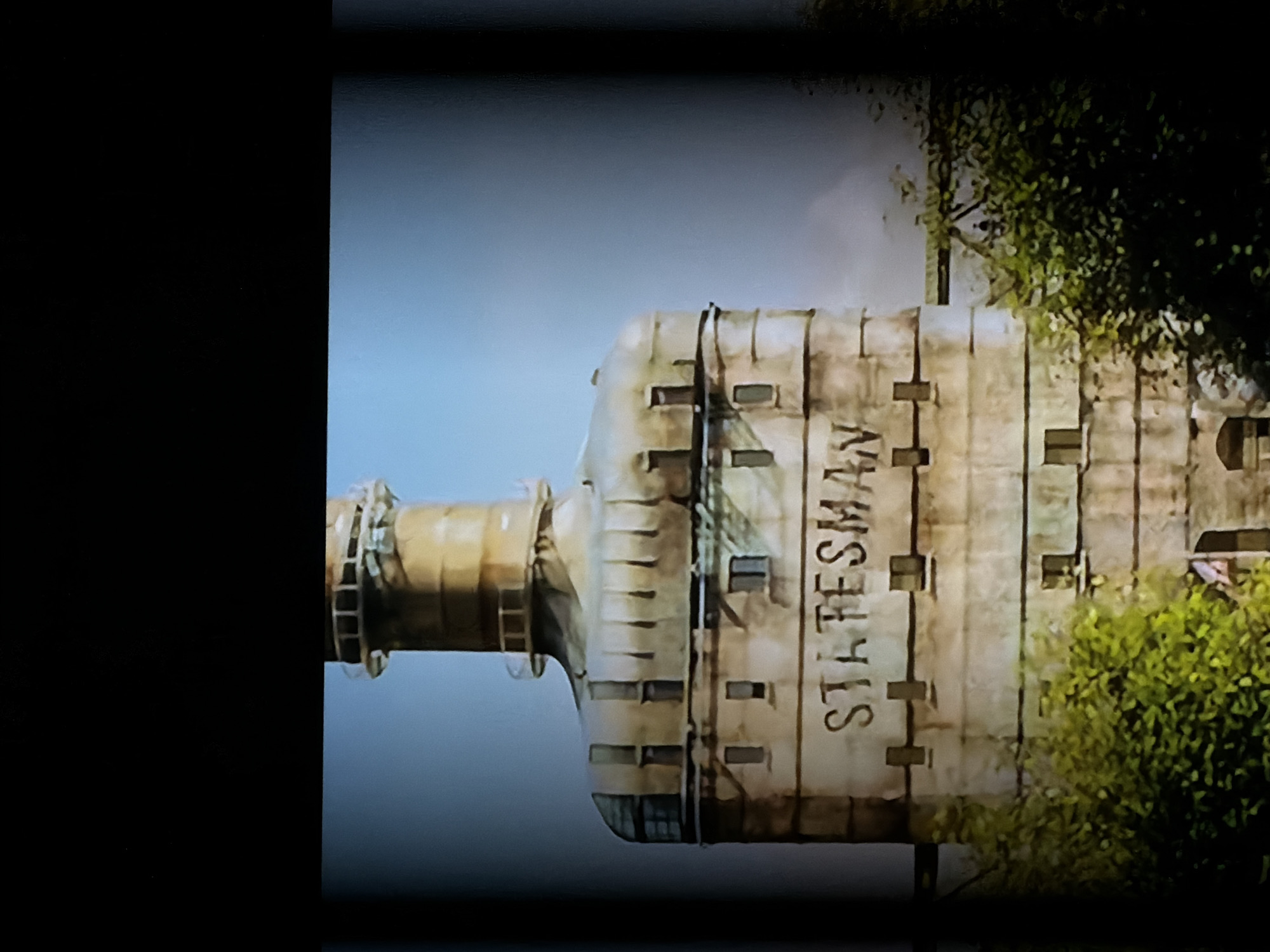The image displays a tall, cylindrical industrial structure, likely a silo or storage tank, set against a clear, light blue sky. The structure features a narrower, pipe-like base that transitions into a wider, multi-segmented body. This main section has a weathered appearance, possibly concrete or metal, with distinct vertical seams and several small, rectangular openings resembling windows or vents. The word "STATESMAN" is prominently painted vertically in large, dark capital letters on the side of the wider structure. Lush green foliage, consisting of bushes and trees, is visible at the base and partially on the right side of the structure. The scene is static, with no human activity visible, suggesting a daytime view in an industrial or agricultural area. The bright sky indicates clear weather. The location context for this structure is Sofiivska Borshchahivka, Ukraine.