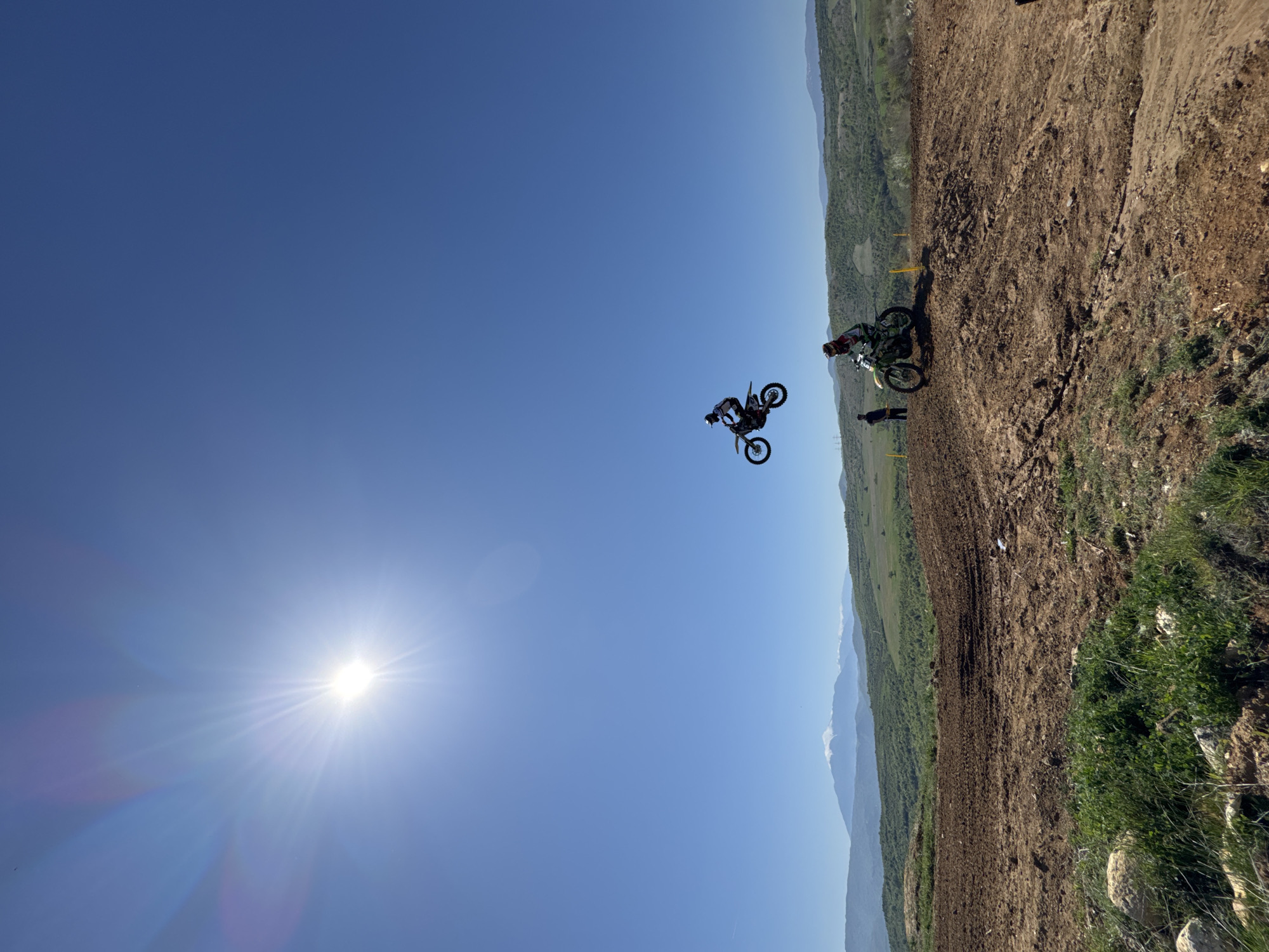 A medium-shot captures a motocross rider mid-air against a bright blue sky with a sun flare. The rider is on a black and green dirt bike, wearing protective gear. The backdrop features a dirt motocross track winding along a grassy, hilly landscape. Another motorcycle and a person can be seen on the track. This scene is at or near Selishte, Bulgaria.
