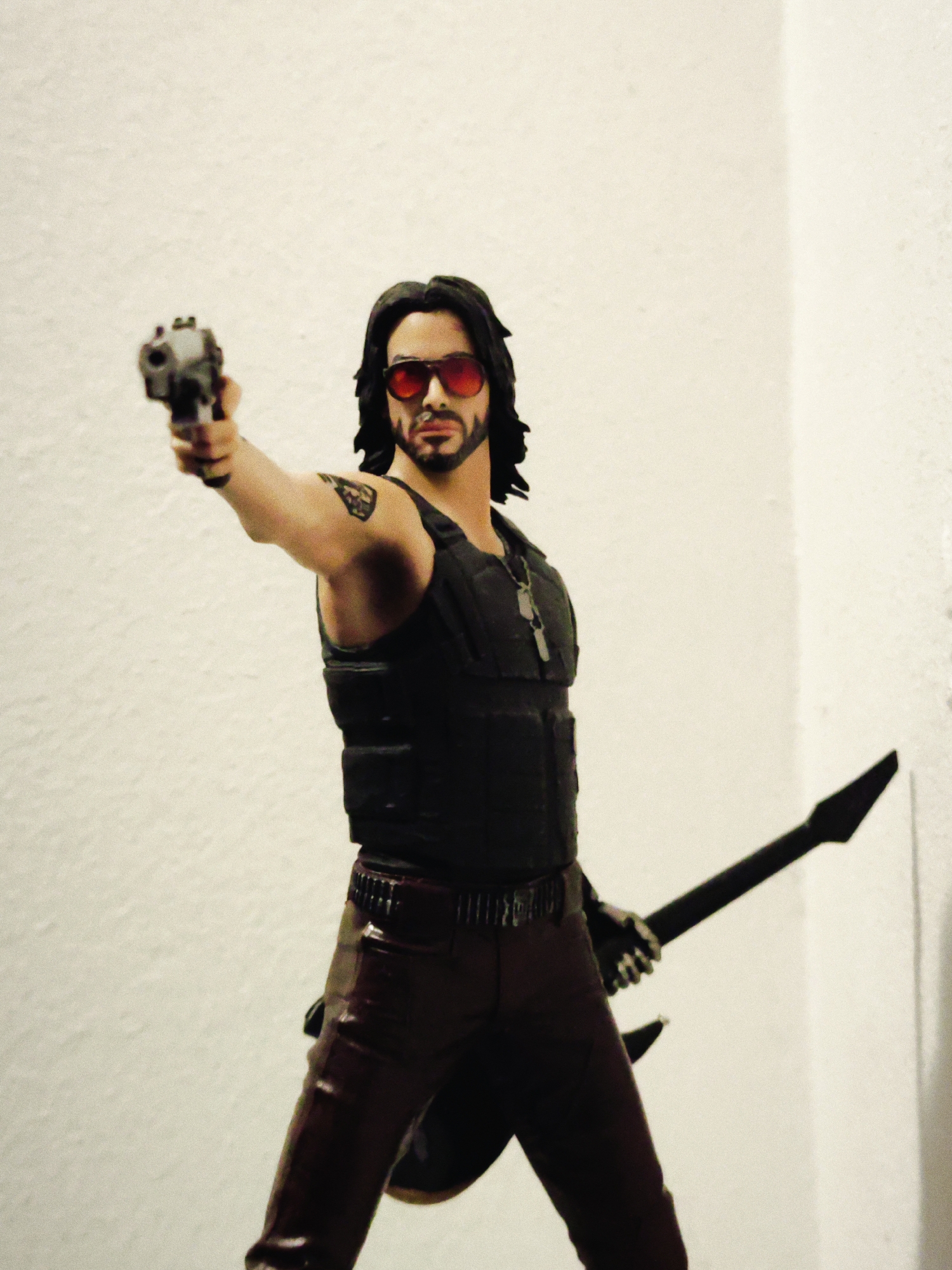 A figurine depicting a man with long dark hair and a beard, wearing red sunglasses, a black vest, brown pants, and holding a gun in his right hand and a black electric guitar in his left hand, stands against a plain white wall. The figurine has a tattoo on its right arm and wears a necklace. The location is not determinable from this image.