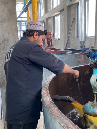 The image shows a man wearing a traditional patterned cap, standing next to a large, circular, concrete tank. He is wearing a black shirt with some text in white on the back. He is holding a stick and appears to be stirring or interacting with the contents of the tank, which contains a viscous, yellow liquid. A pipe, wrapped with a black material, protrudes into the tank. Other machinery and pipes are visible in the background, suggesting an industrial setting. Windows are visible in the distance, providing some natural light. The location is likely in the city of Kandahar, Afghanistan, based on the cultural attire of the man.