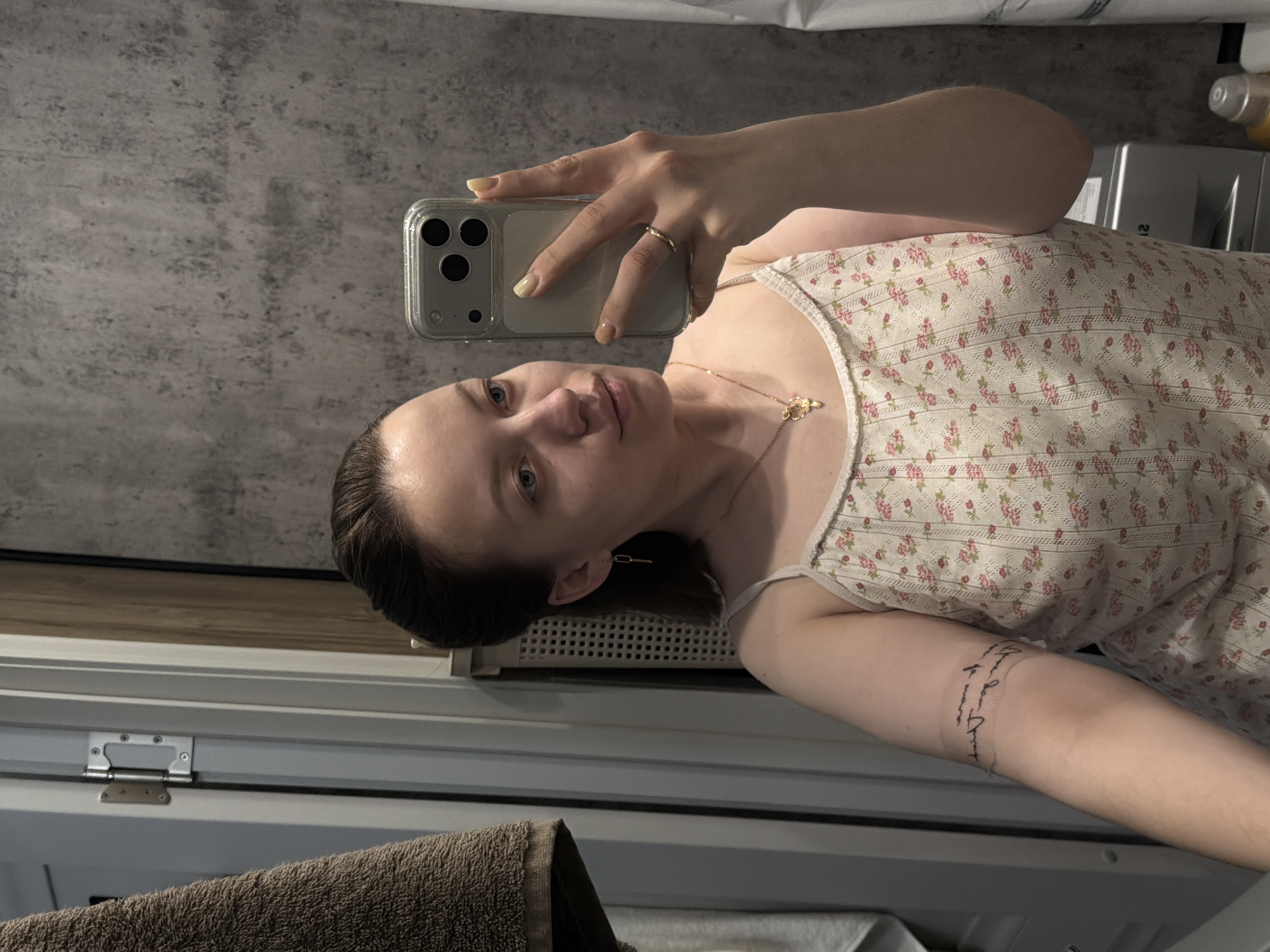 A woman is taking a selfie in a bathroom. She is wearing a floral patterned top and a gold necklace. She has a tattoo on her arm. The bathroom has gray walls and a white cabinet. There is a towel visible in the lower left corner. There is no information indicating that the location is Reshetnikovo, Russia.