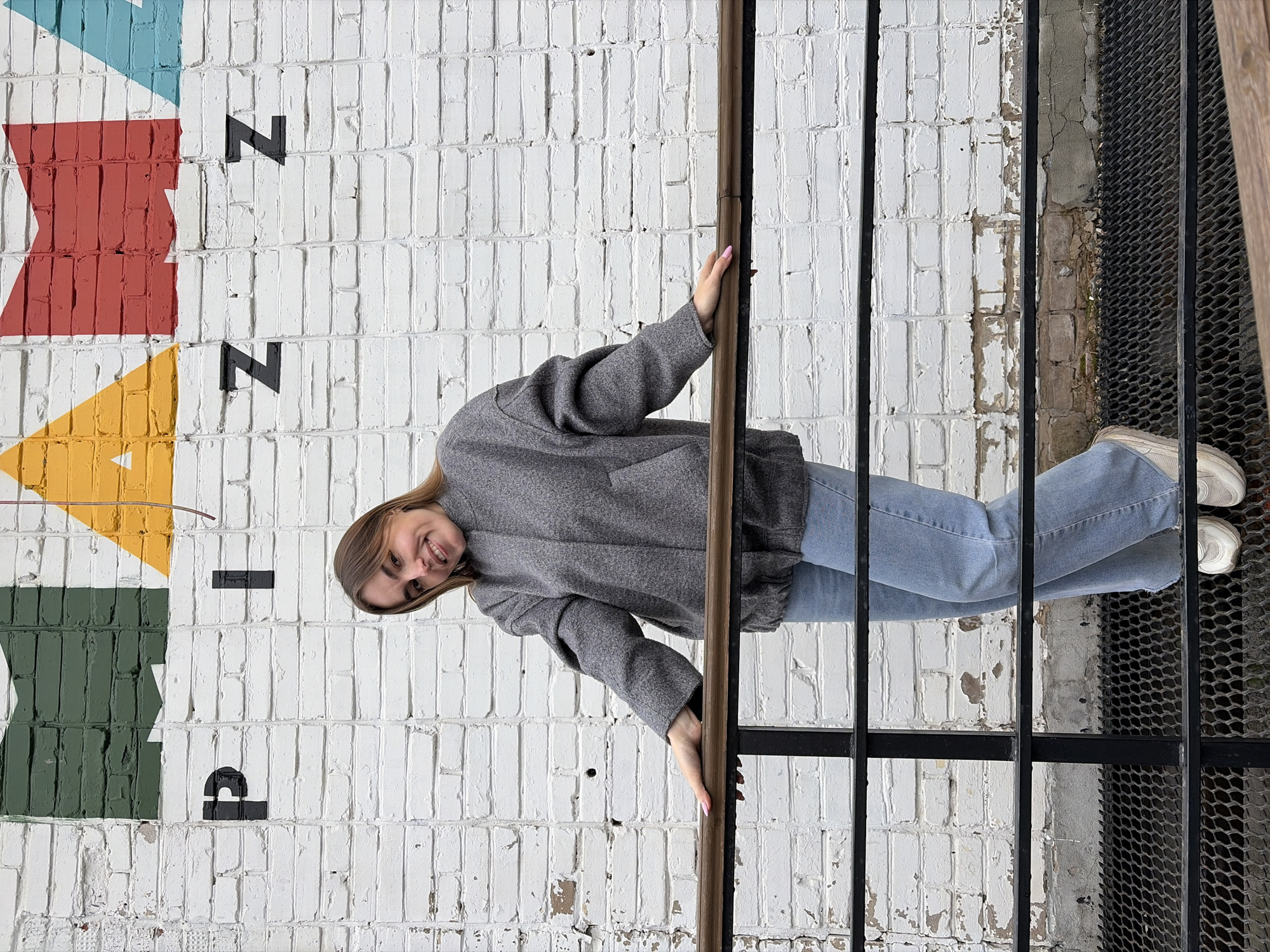 A woman is standing in front of a painted brick wall in Izhevsk, Russia. The wall has geometric shapes painted in red, yellow, and green, and the letter "Z" is painted in black. The woman is wearing a gray coat and blue jeans, and white sneakers. She is holding onto a metal railing. The location is a public art installation in Izhevsk.