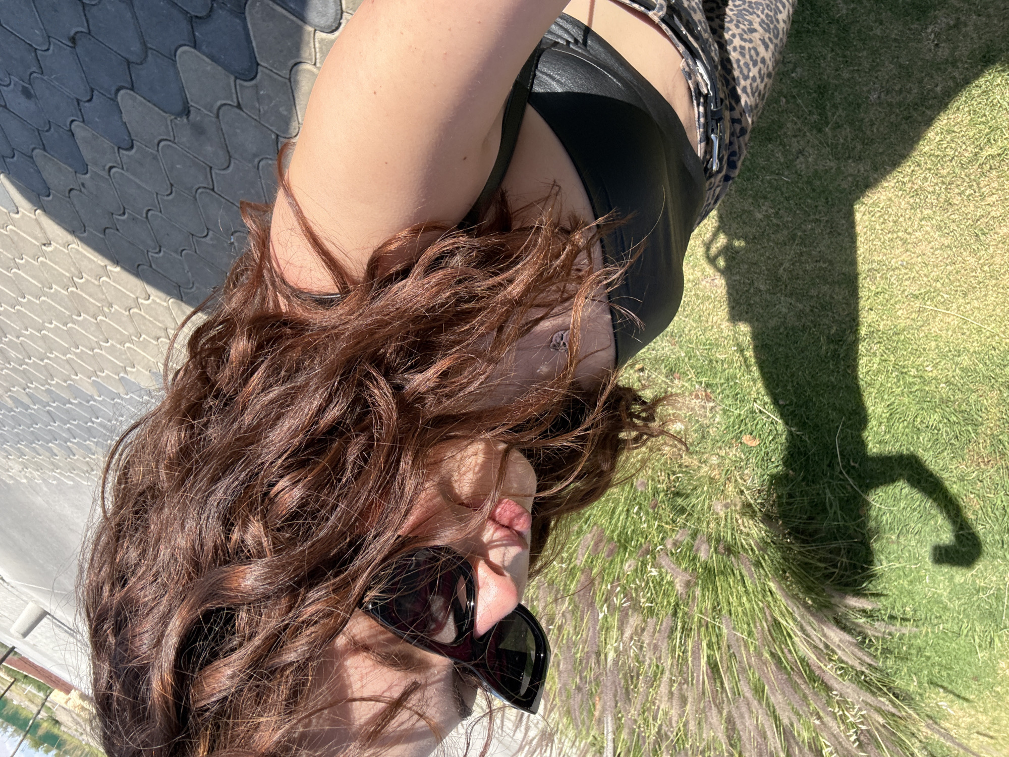 A woman with long, curly, reddish-brown hair is lying on her side in the grass, wearing sunglasses, a black top, and a leopard-print belt. Part of her body is on a patterned pavement. The image appears to be taken in daylight, with the sun casting a shadow on the grass. The background includes some greenery and a distant view of a building and water. There is no verifiable way to locate the exact spot within Santa Clara Ocoyucan from the image.