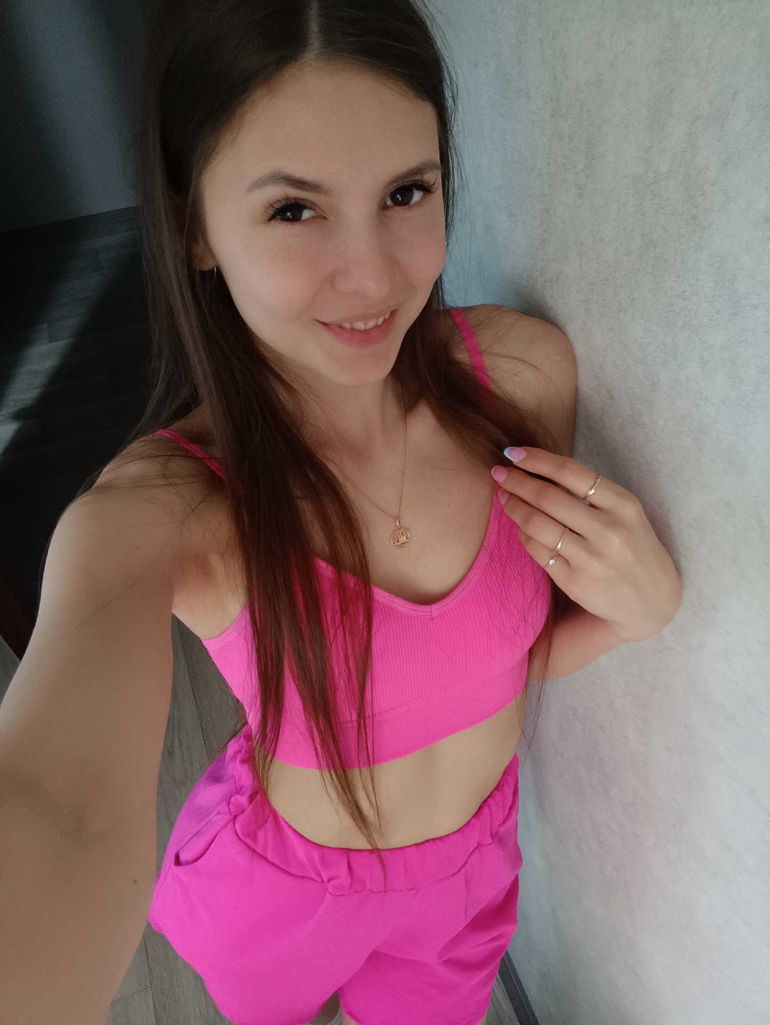A young woman poses for a selfie in an indoor environment. She is wearing a bright pink crop top and matching shorts. A gold necklace adorns her neck, and rings are visible on her fingers. She is standing against a textured, light-colored wall. To the left, there is a glimpse of a dark-colored wall or doorway. The floor appears to be wood-toned. The woman is smiling slightly and looking directly at the camera. The location is Unknown, Unknown.