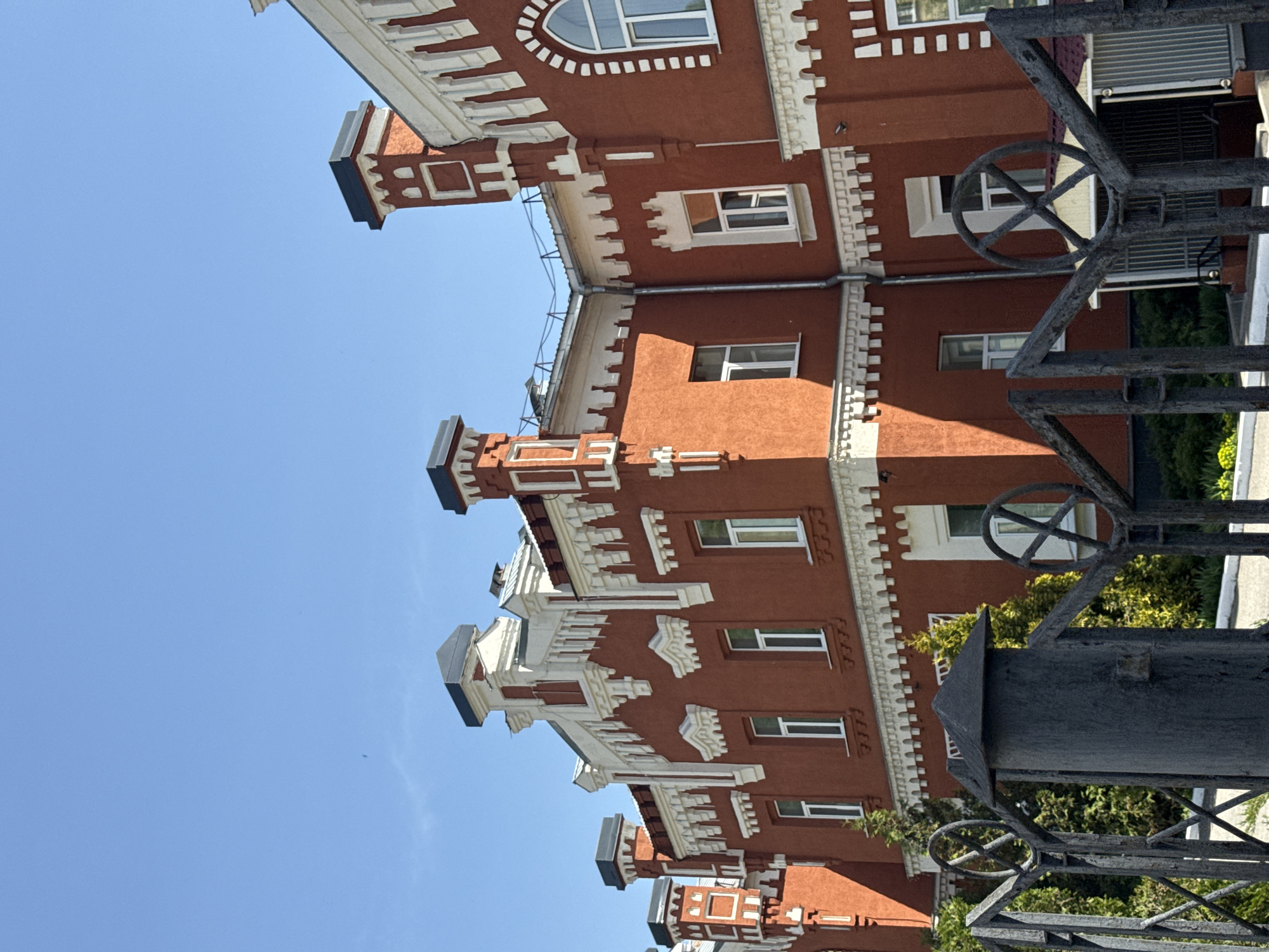 The image depicts a multi-story building with red brick walls and white architectural details, likely located in Penza, Russia. The building has numerous windows of varying sizes and shapes, some with arched tops. The architectural style appears to be historic, potentially from the late 19th or early 20th century. A wrought-iron fence with decorative elements, including circular motifs, partially obscures the view of the building. The sky is a clear, bright blue. Green foliage is visible behind the fence, suggesting a garden or park nearby. The image is oriented vertically, with the building appearing to extend upwards into the sky.
