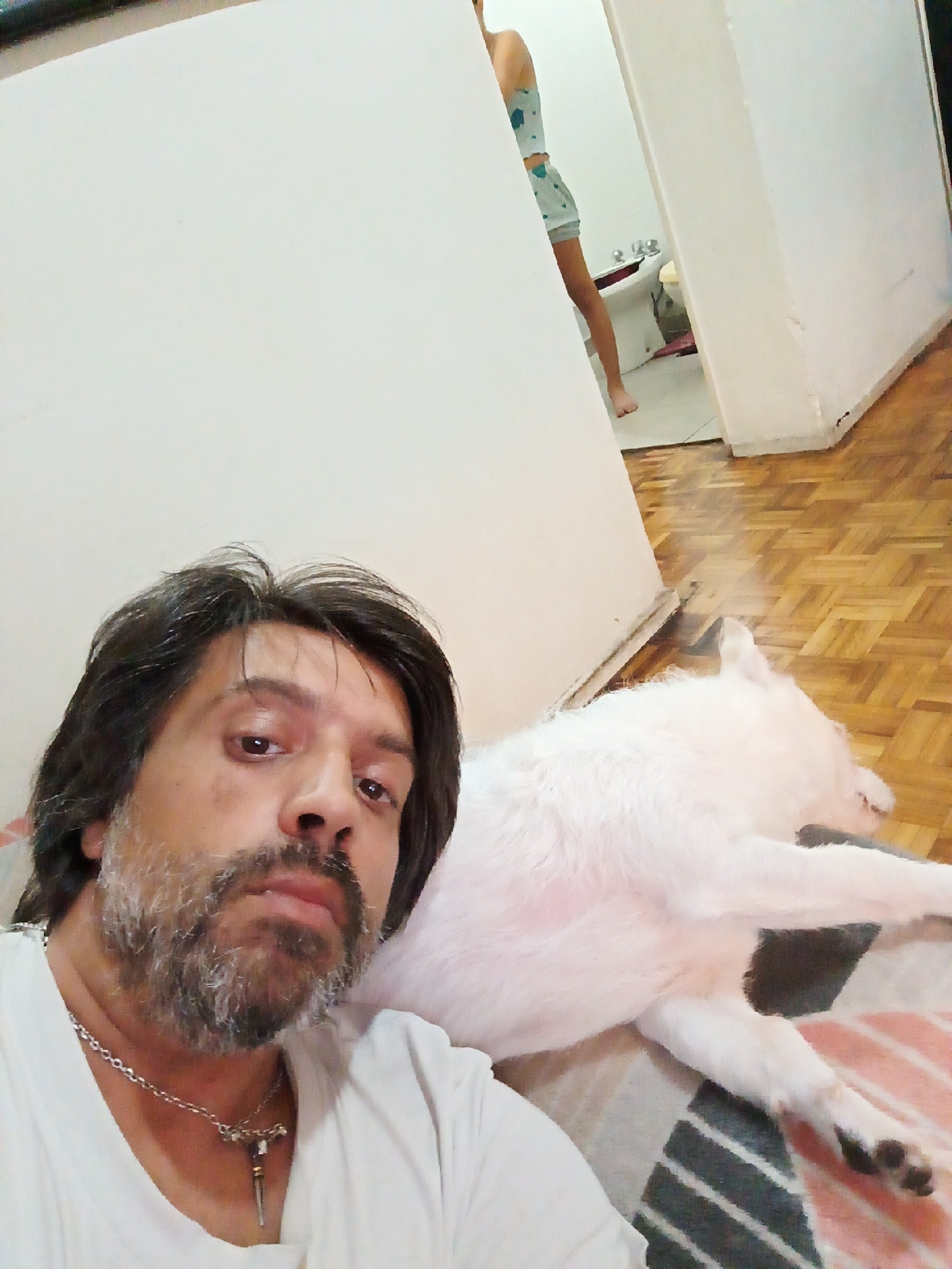 A selfie taken by a man with dark hair and a grey beard, wearing a white t-shirt and a silver chain with a pendant. He is lying down next to a white dog. In the background, through a doorway, a person wearing a light blue and white patterned top and shorts is visible in a bathroom. The floor is parquet. The location is likely a home in Buenos Aires, Argentina.