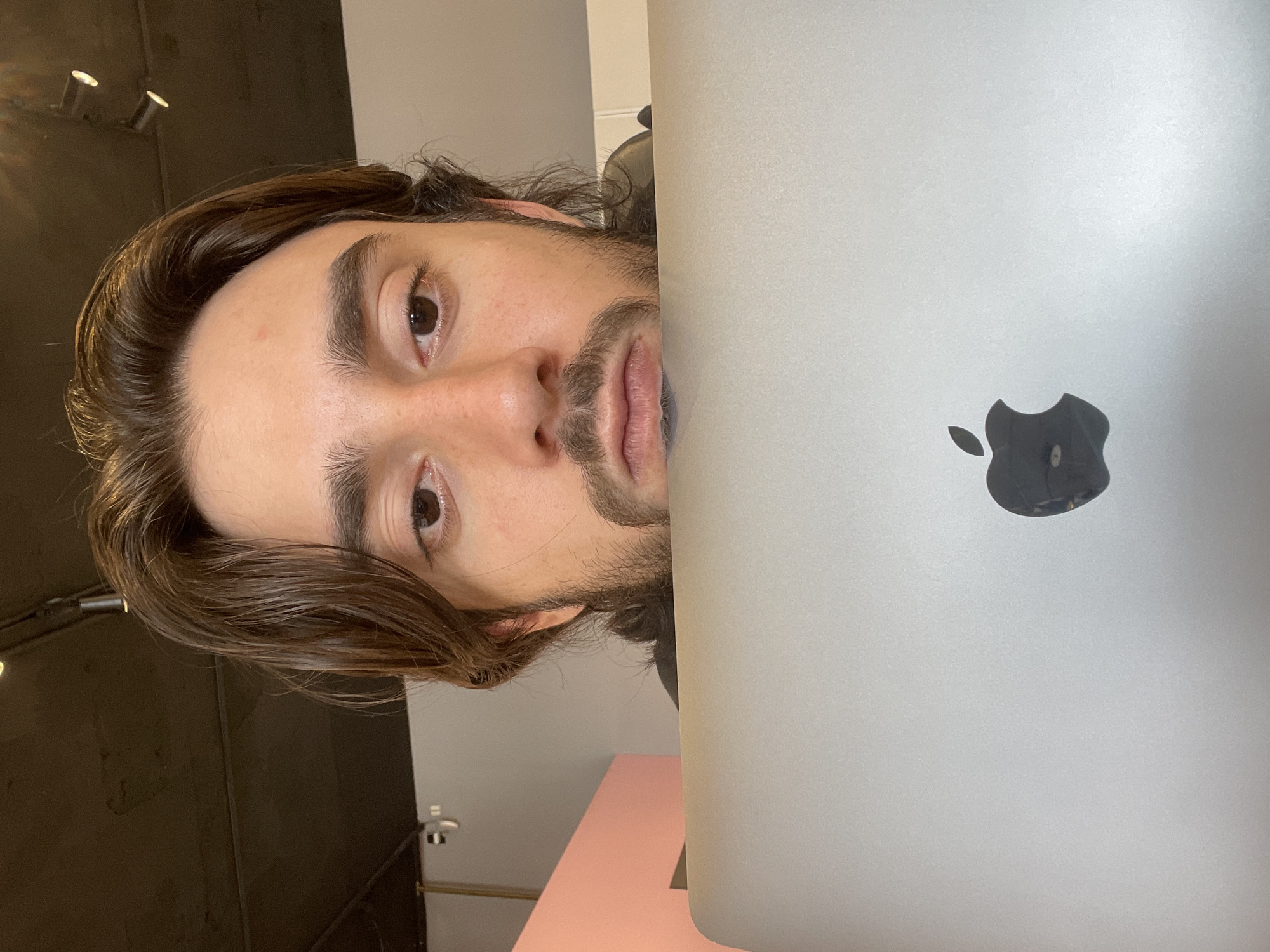 A selfie of a man with long brown hair, brown eyes, and a beard, taken indoors in Mexico City, Mexico. The man's face is partially obscured by a silver Apple laptop. The background shows a black wall with track lighting, a white wall, and a pink table. A small security camera is visible on the white wall.