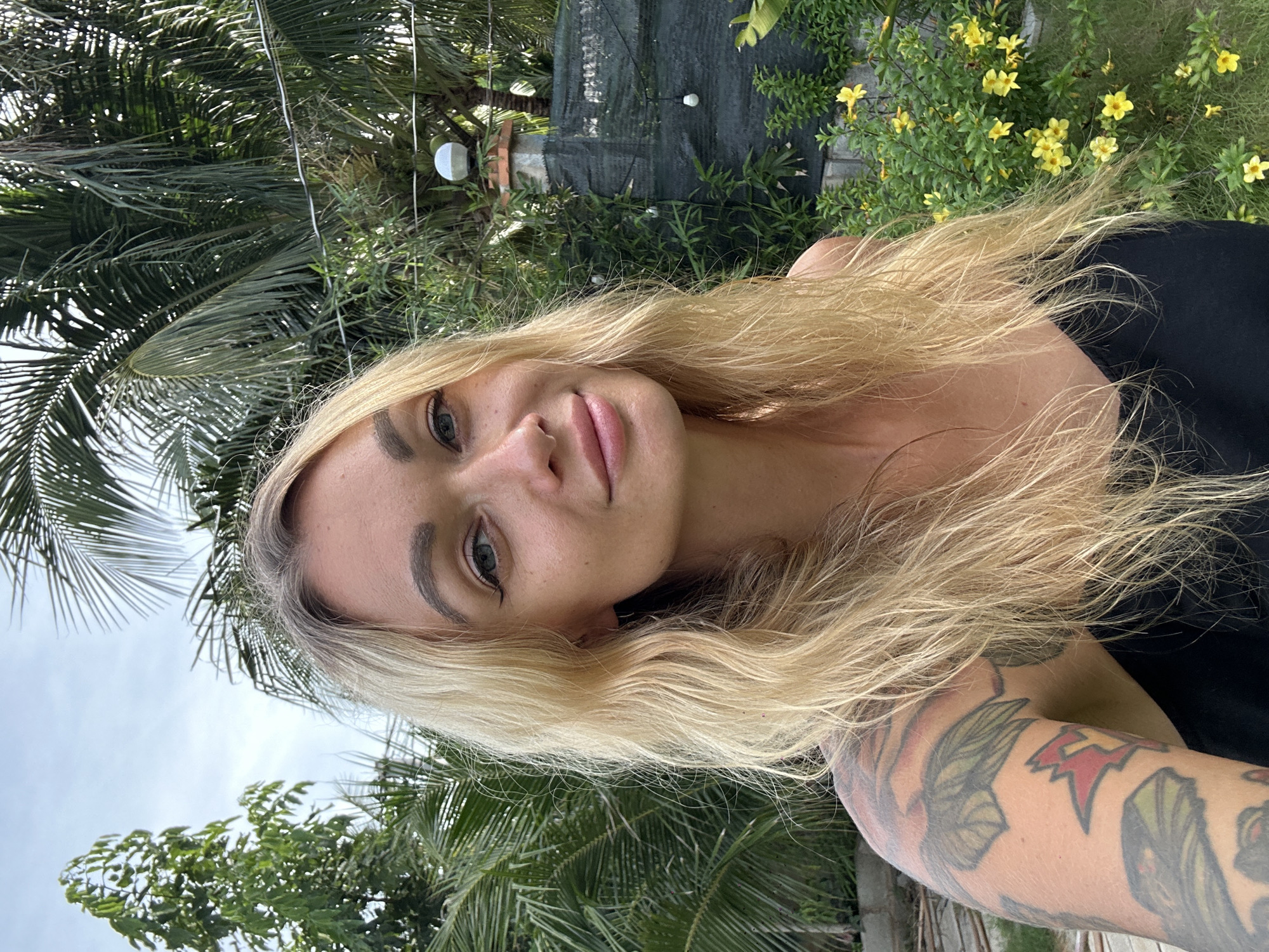 This media file shows a close-up, vertical selfie of a blonde woman outdoors. She is positioned in the foreground, with her face tilted towards the camera, smiling subtly. Her long, wavy blonde hair frames her face. She is wearing a black top. Her left arm, visible on the right side of the frame, is heavily tattooed with colorful designs, including what appear to be wings and geometric shapes.
The background suggests a tropical environment, with lush green palm fronds and foliage dominating the scene. In the upper background, a bush with small yellow flowers is visible. There are also some subtle hints of man-made structures, including what looks like a dark mesh screen and possibly some brickwork. Two reflective spherical objects, possibly garden ornaments, are also present in the upper left portion of the background, one catching the light. The sky is overcast, suggesting it is daytime, possibly late afternoon or early morning given the soft lighting. The overall impression is one of a relaxed, outdoor setting in a warm climate. The location context provided is Mũi Né, Vietnam, which aligns with the visual cues of tropical vegetation. No text is visible in the image.