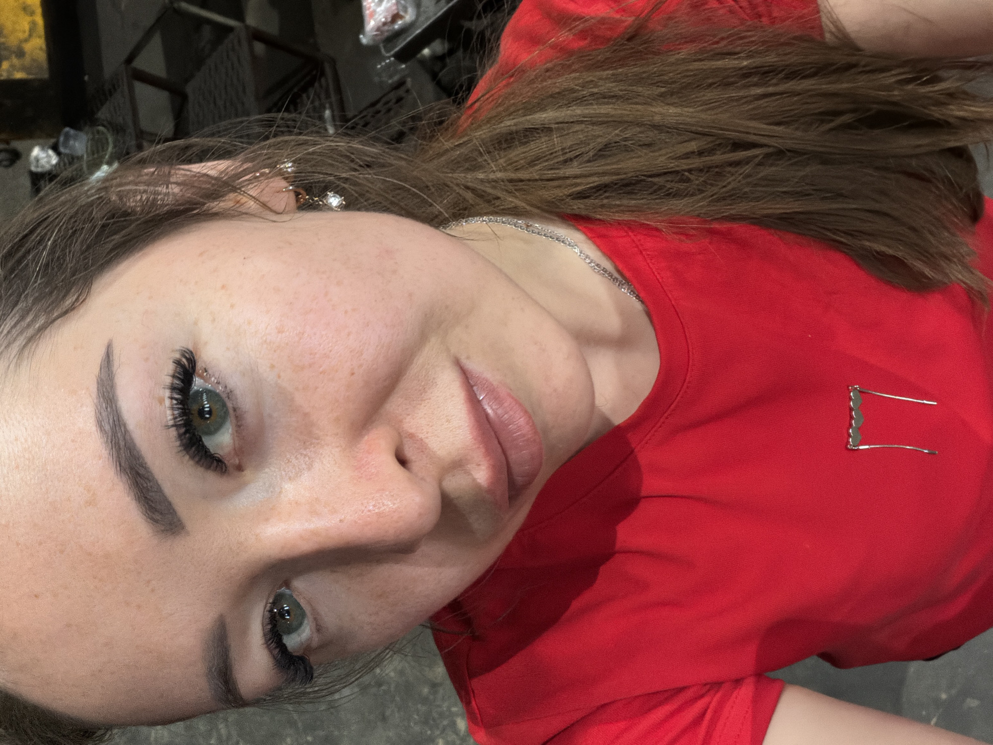 A woman with light brown hair and green eyes is pictured in a close-up, high-angle shot. She is wearing a red t-shirt adorned with a heart-shaped brooch and a silver chain necklace. Her makeup includes dramatic false eyelashes and subtle lip gloss. She has numerous freckles across her face and décolletage. The background is blurred, showing dark industrial shelving and what appears to be a concrete floor. The image seems to be taken indoors. There is no indication of the specific location within the city of Oktyabrsky, Russia.