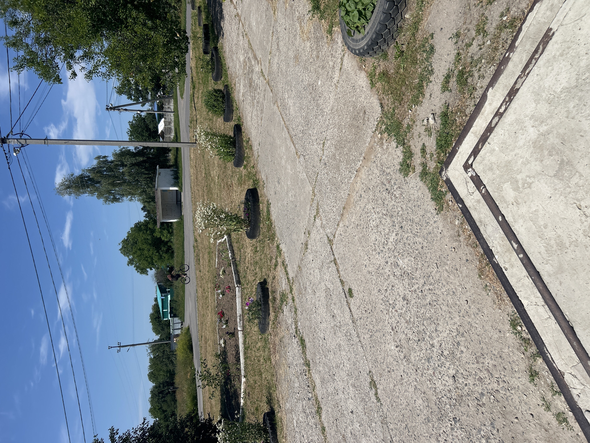 A person is cycling on a road in Malyi Trostianets, Ukraine. The road is paved and a few buildings are visible in the background, along with trees and a power line pole. There is a flowerbed in the foreground, decorated with flowers and some tires used as planters. The sky is blue with scattered clouds.