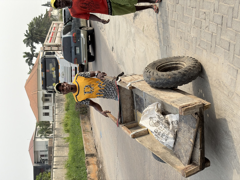 A man smiles and pulls a wooden cart with a tire on top down a street in a residential area. Another man stands nearby. Behind them, a line of vehicles waits on the street, including a gray SUV and a large white truck. A bag is visible in the cart, and houses line the street. The sky is clear, and the sun is shining.
