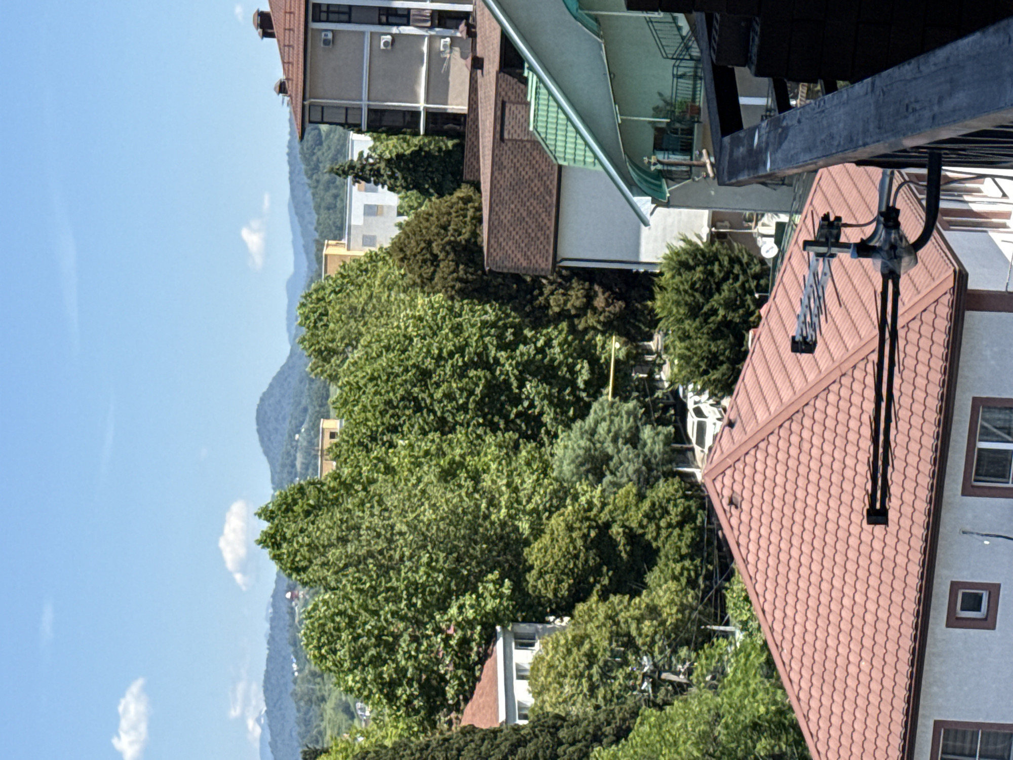 The image captures a view of urban and natural landscapes in Sochi, Russia. In the foreground, a building with a red tiled roof is visible, featuring white walls and windows. An outdoor television antenna is mounted on this roof. Adjacent to it is another building with a light-colored facade and windows. Further back, a larger building with a dark tiled roof and multiple windows is present. Behind these structures, lush green trees dominate the scene, interspersed with other buildings of varying heights and architectural styles. In the distance, a range of rolling hills and mountains, covered in vegetation, forms the backdrop. The sky is blue with scattered white clouds, indicating a clear day. The overall impression is one of a coastal city surrounded by natural beauty, with a blend of residential and possibly commercial buildings. There are no people visible in the image.