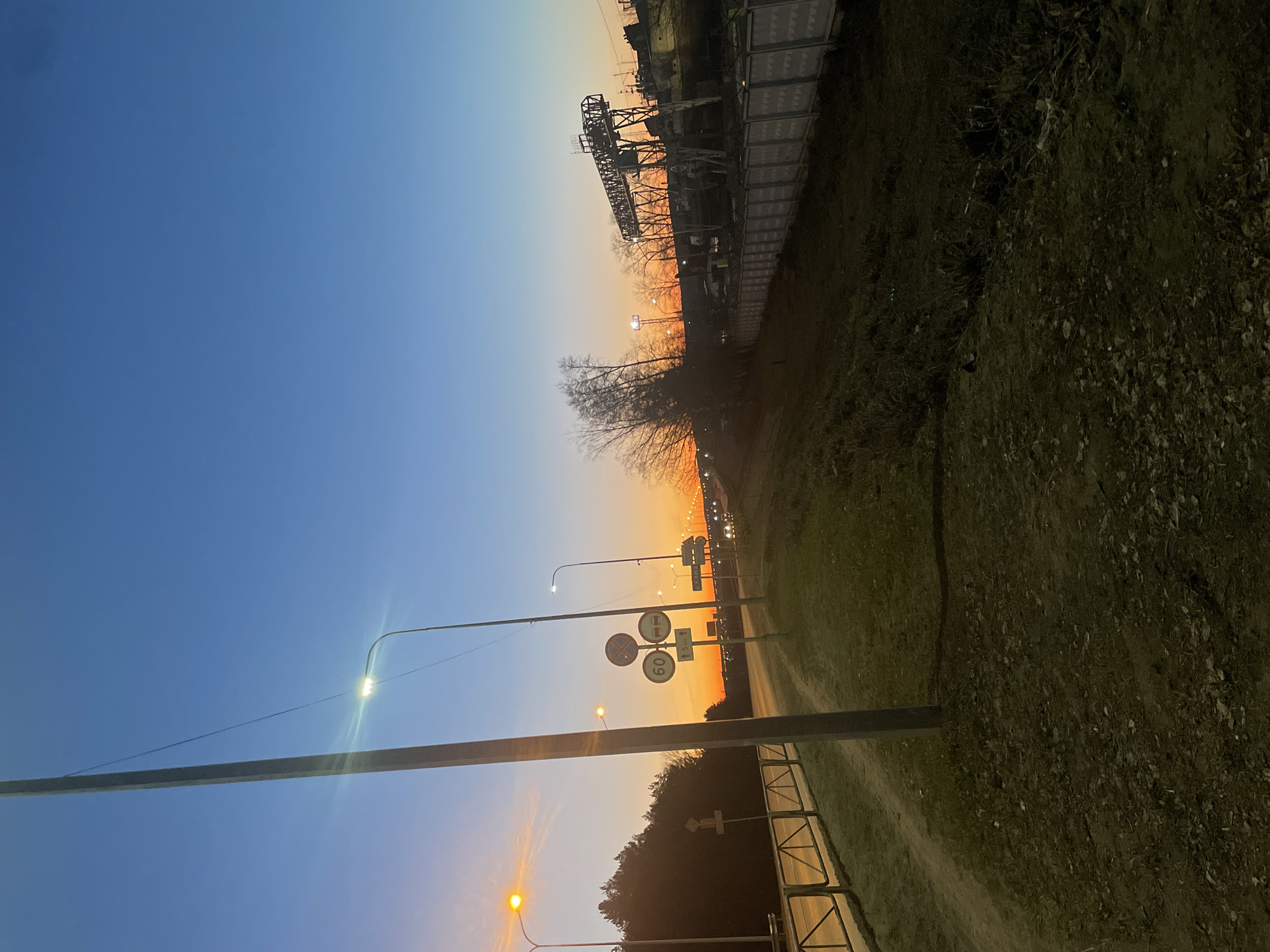 This image shows a street scene in Chaikovsky, Russia, during dusk or dawn. The sky transitions from blue to orange near the horizon. Streetlights illuminate the scene. A bridge or elevated structure is visible in the background. There are road signs. Vegetation, including trees and grass, lines the road. An industrial structure is at the top of the image.
