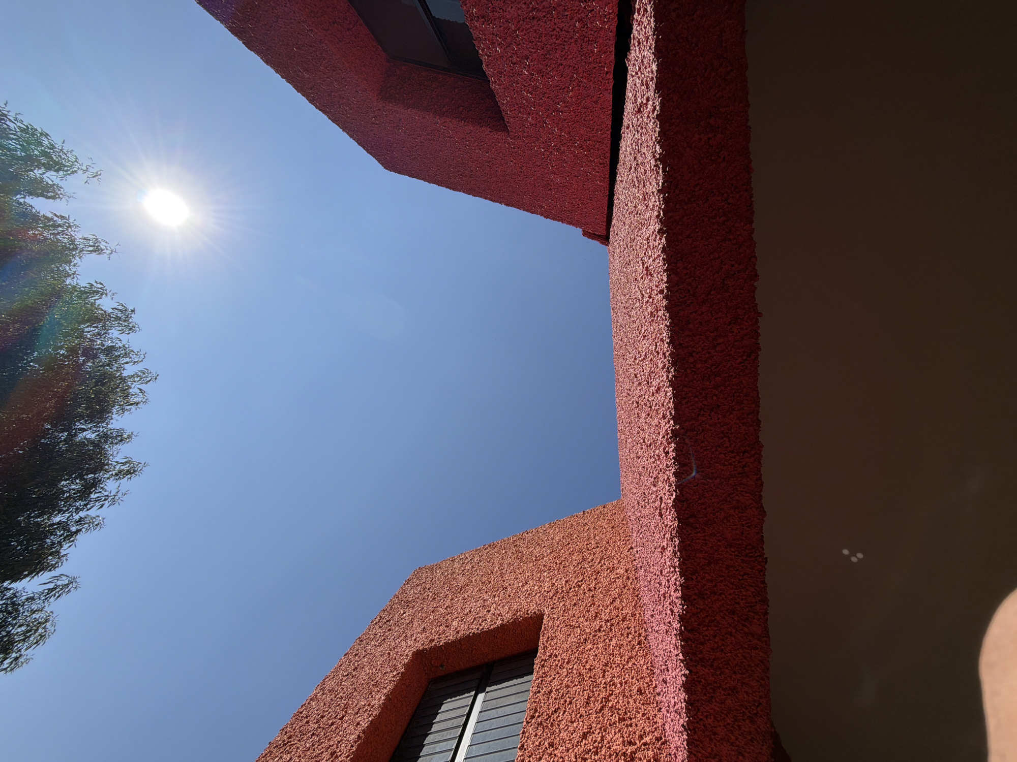 The image, taken from below, depicts the exterior of a reddish-orange building with a rough, textured surface. A tree with dark foliage is visible to the left, positioned against a bright blue sky with the sun shining intensely. The building has what appears to be windows with horizontal blinds. The location is San Andrés Cholula, Mexico.
