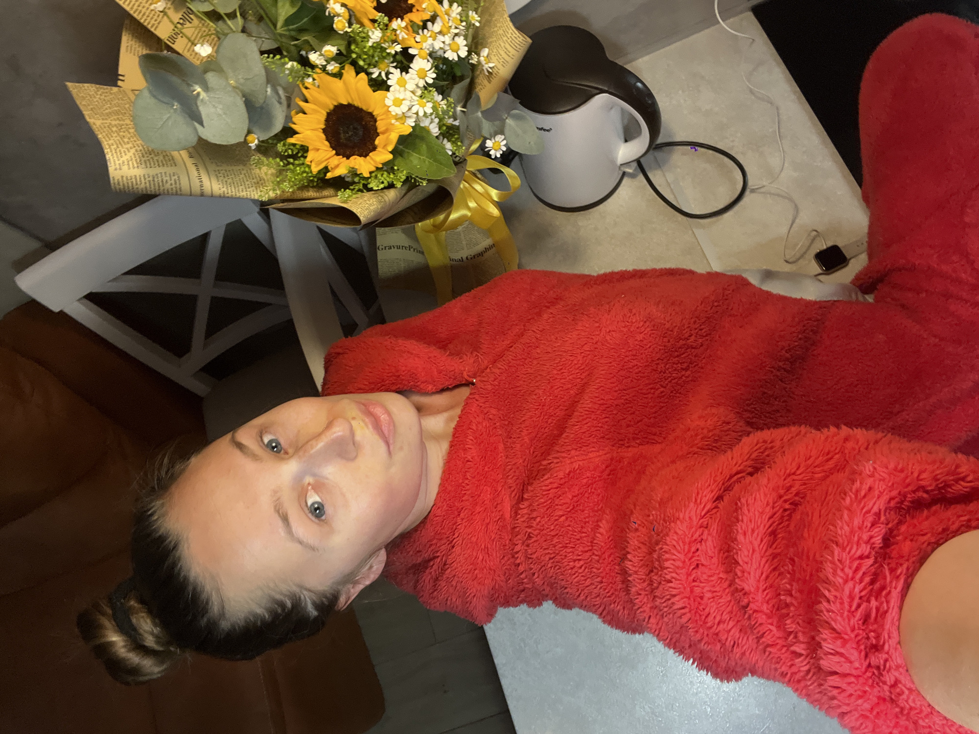 The image depicts an indoor scene in Bečići, Montenegro. A female subject, appearing to be an adult, with light skin, blue eyes, and dark hair pulled into a high bun, is shown in a close-up, upward-angled shot. She wears a red, fuzzy hooded garment and gazes directly at the viewer with a slight pout; a small mark or indentation is visible on her right nostril. To her right, on a light-toned table surface, a bouquet is positioned, containing sunflowers, white daisies, and green eucalyptus foliage. The bouquet is wrapped in brown paper with newspaper-like text and tied with a yellow ribbon. Beside the bouquet, a white and dark grey electric kettle with an attached black cord is visible. The background reveals a dark upholstered sofa and a white chair with a distinctive cross-brace backrest. The lower right portion of the image shows a segment of a light-colored floor, on which a dark square object, resembling a smartwatch, and additional cables are present.