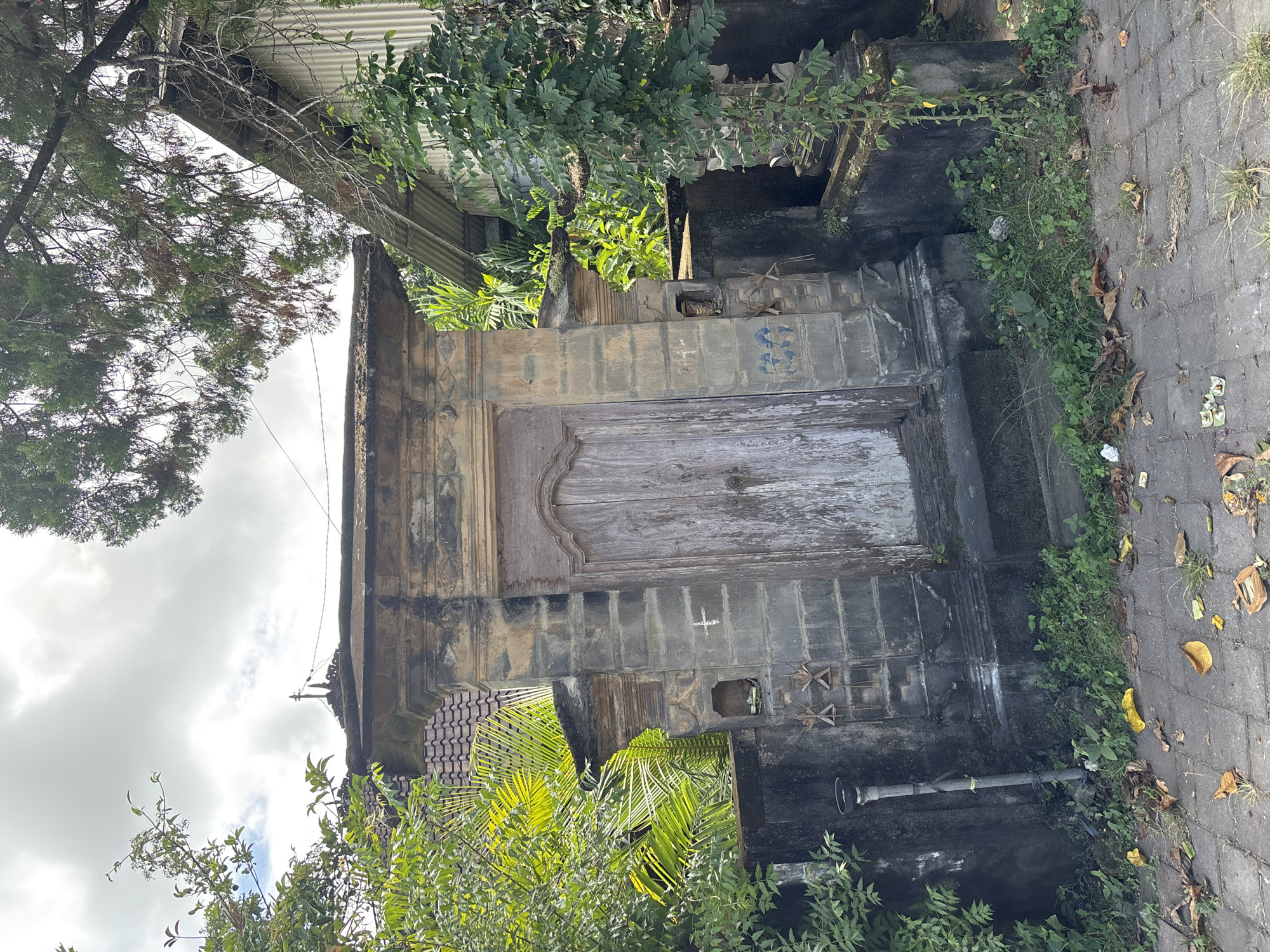 An outdoor view of a deteriorated building in Kuta Selatan, Indonesia. The building has an aged, wooden door, set into a dark, possibly concrete or stone frame. The structure is flanked by lush green vegetation on either side, with trees visible above and below. A portion of a brick-paved surface is visible on the right side. The building appears to be in a state of disrepair, with visible weathering and possible water damage.