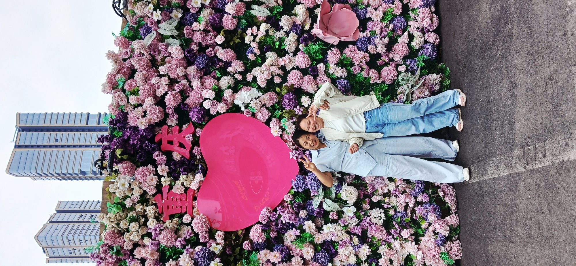 Two women lie side-by-side on a flower wall. One woman wears a white jacket with blue jeans. The other wears a light-blue shirt and pants. Both women flash peace signs. The flower wall contains pink, purple, and white flowers. A large, pink heart-shaped sign rests near them on the flower wall. Chinese characters also appear on the wall. In the background, a tall building is visible, suggesting an urban setting. The scene is located in Yuzhong District, China.