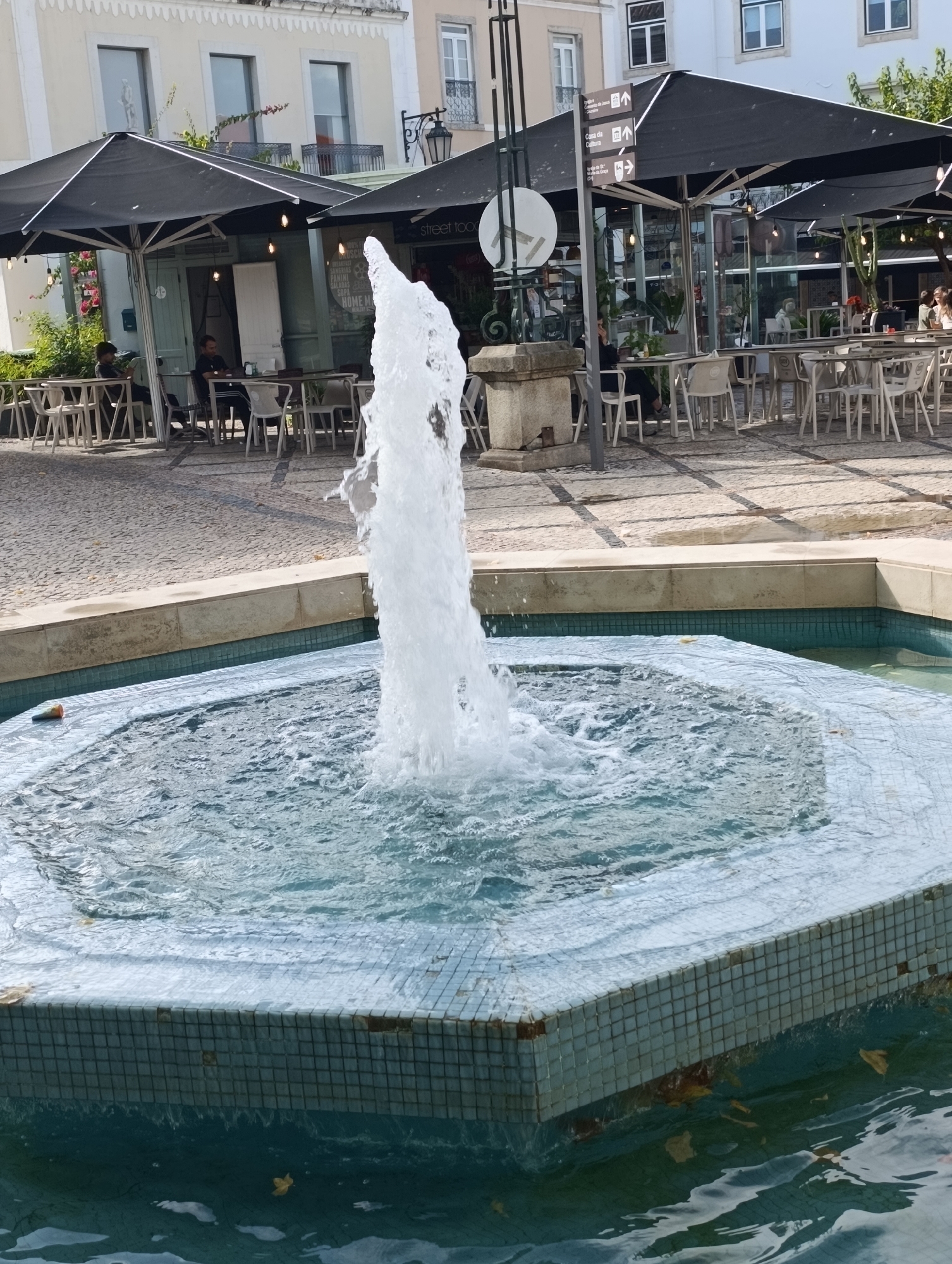 A polygonal fountain with a tiled edge is in the foreground, with a powerful jet of water erupting from its center. The water in the fountain is clear and reflects the sky. Beyond the fountain, a cobblestone plaza is visible, with several outdoor seating areas and black umbrellas providing shade. Restaurants and buildings with multiple windows line the plaza. In the background, a white building with a distinctive patterned cornice stands out. People are seated at tables, some dining and others engaged in conversation. A signpost with directional arrows is also present. The overall scene depicts a lively outdoor public space, likely a city square or promenade, suggesting a warm, pleasant day. The specific location within Setúbal, Portugal is likely Praça do Bocage or a similar central square given the architectural style and presence of outdoor dining.
