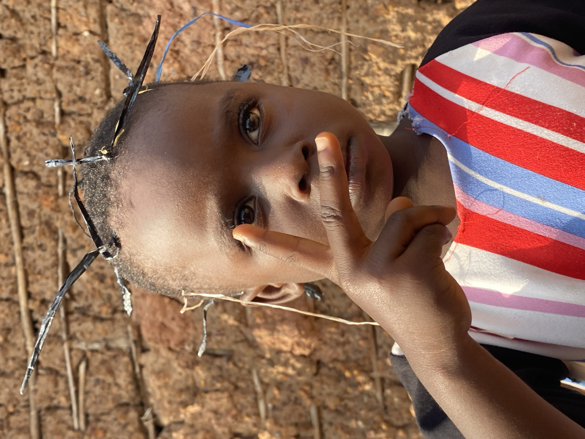 A young child of African descent stands centered in the frame, facing the camera directly. The child has dark skin, short dark hair, and brown eyes. Their head is adorned with several thin, dark, metallic-looking objects, appearing to be attached to the hair. The child's right hand is raised, with the index and middle fingers extended and touching their lips, as if in thought or to quiet themselves. The child is wearing a shirt with a black base and a brightly colored striped pattern of red, white, blue, and pink across the chest. The background consists of a textured, reddish-brown wall, possibly made of mud or brick, with visible horizontal and vertical elements. The lighting suggests it is daytime, casting a slight shadow on the child's face. The overall impression is a portrait captured outdoors, with a rustic environment suggested by the background. This image was captured in Ufu Esioni, Nigeria.