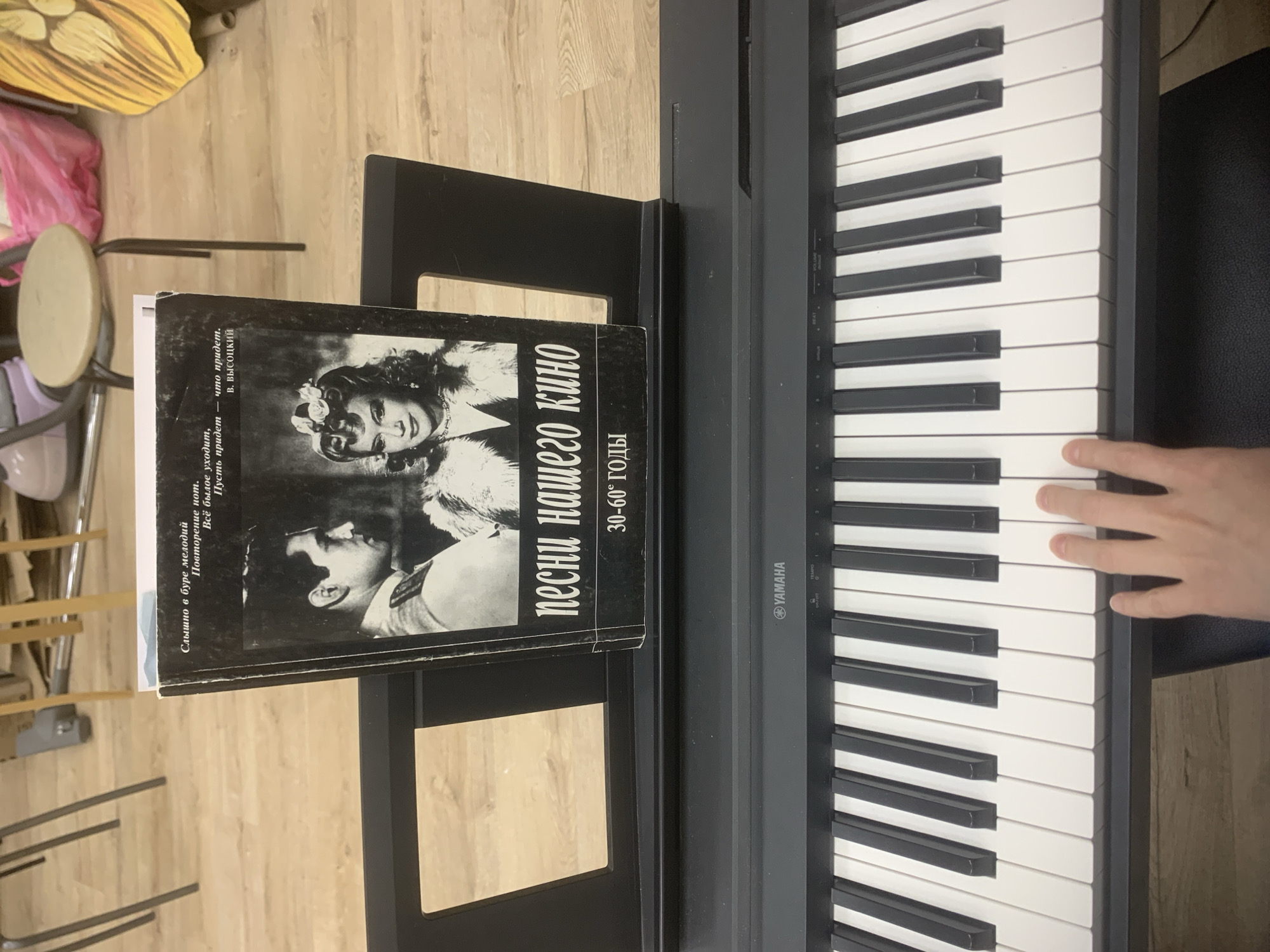 This vertically oriented image captures an indoor scene, likely a music practice space, in Izhevsk, Russia. The focal point is a black Yamaha digital piano, with a person's right hand positioned over the white keys, suggesting they are either playing or about to play. A black music stand is attached to the piano, holding a Russian book.
The book's cover features black and white vintage photographs, possibly film stills, and prominent text in Cyrillic. The main title reads "ПЕСНИ НАШЕГО КИНО" (Songs of Our Cinema) and indicates "30-60 ГОДЫ" (30s-60s), suggesting a collection of songs from Russian cinema of that era. Text visible on the book's spine or side includes a quote attributed to "В. ВЫСОЦКИЙ" (V. Vysotsky): "Слышно в буре мелодий / Повторение нот. / Всё былое уходит, / Пусть придет что придет." (Heard in the storm of melodies / Repetition of notes. / All the past departs, / Let come what may.)
The digital piano displays the "YAMAHA" logo and partially visible controls for "VOLUME" and "TEMPO". The floor is made of light-colored wooden planks. In the background, various objects are discernible, including a tan circular object resembling a drum or stool, part of a pink bag, and a yellow-orange decorative item, contributing to the casual and lived-in atmosphere of the setting. The scene depicts an individual engaged in musical practice or enjoyment.